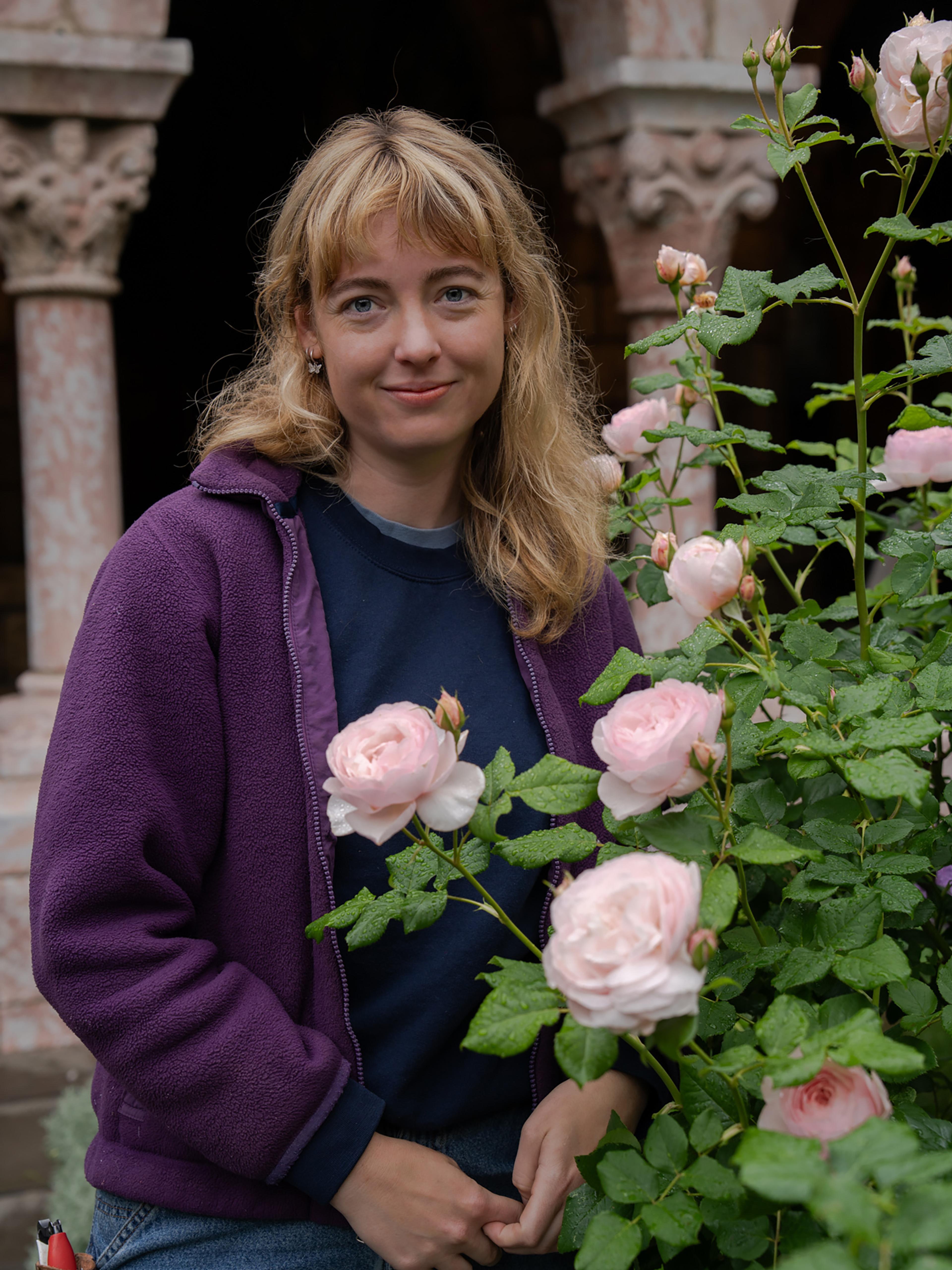 A woman with blonde hair smiles behind a bush of pink roses