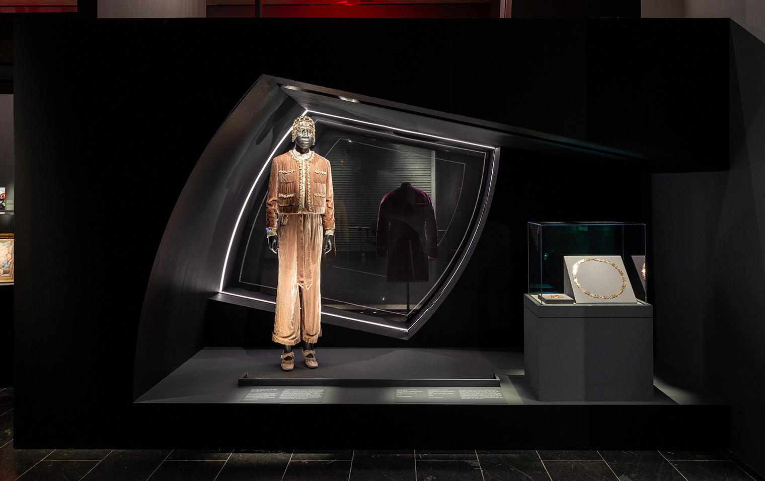 A mannequin in a beige, ornate jumpsuit with headpiece in a futuristic black display. Nearby, a gold necklace is showcased under soft lighting.