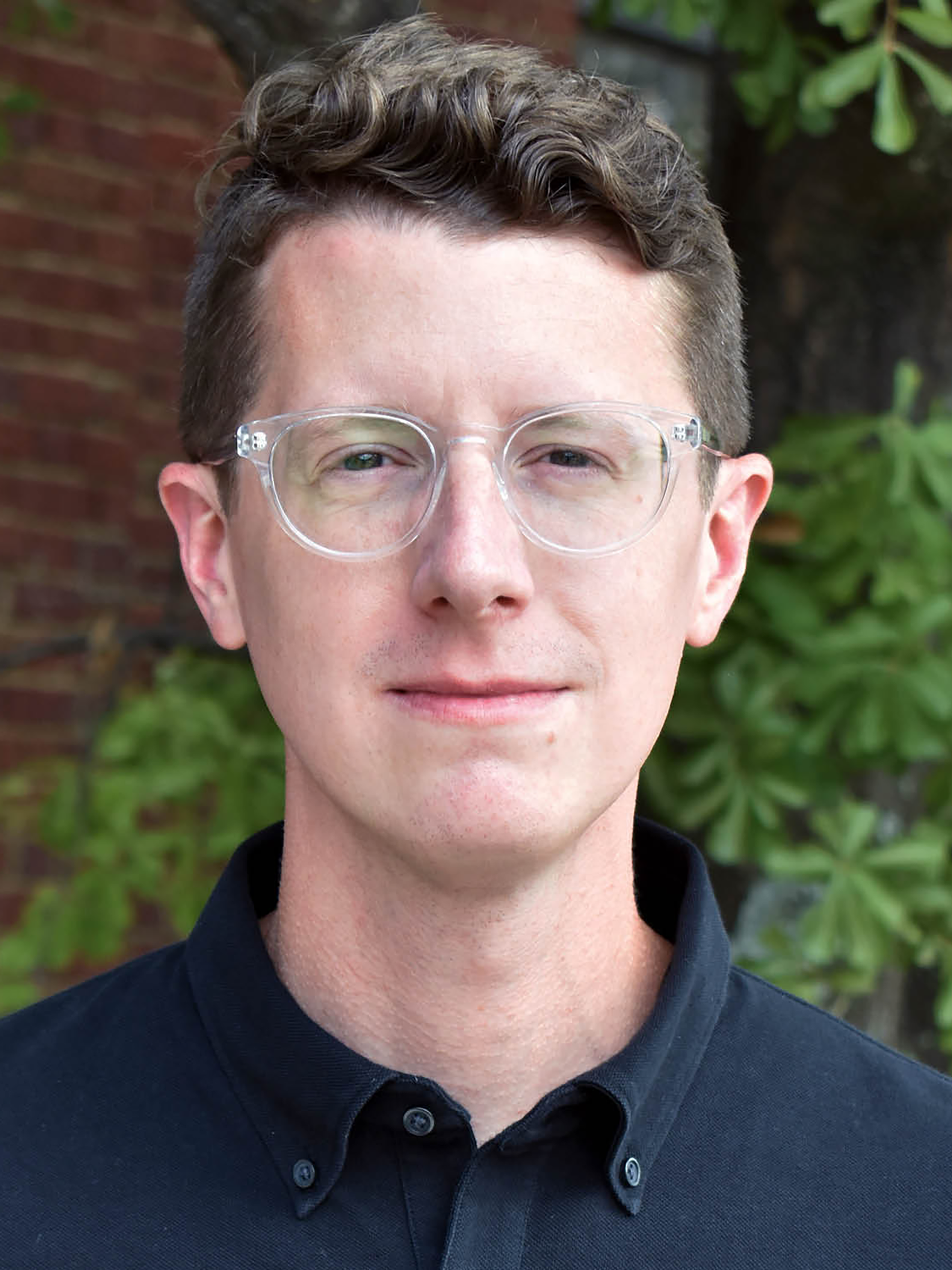 A man wearing glasses stands in front of a brick building