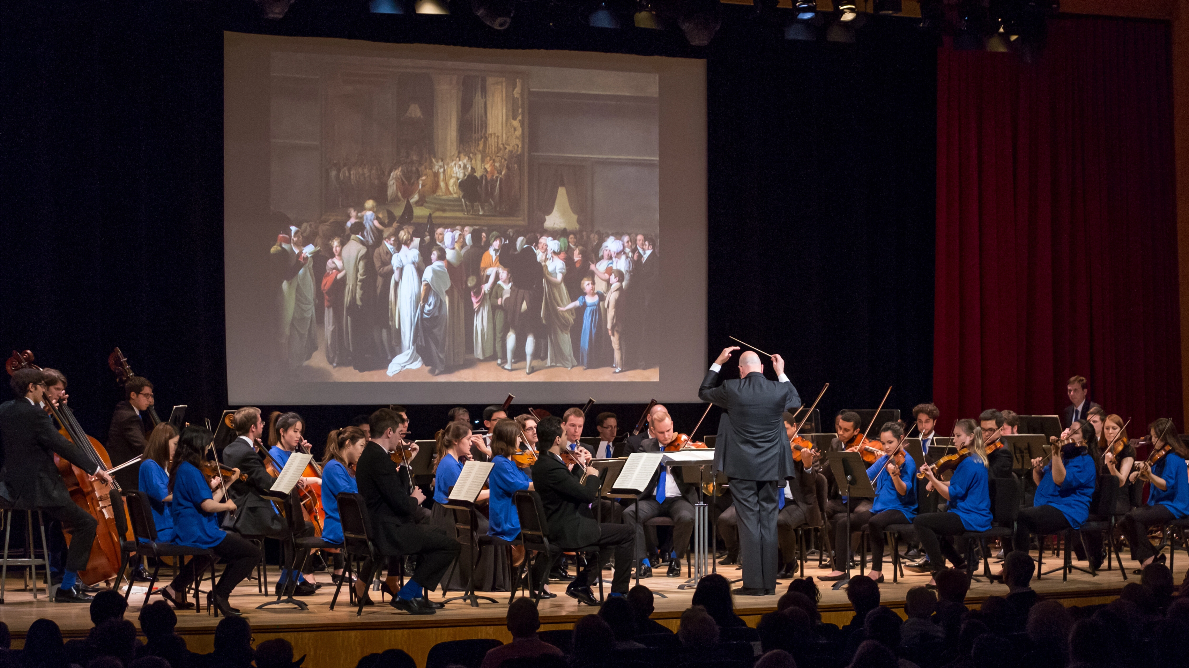 An orchestra performs on stage. An artwork shows on a projector screen above them.