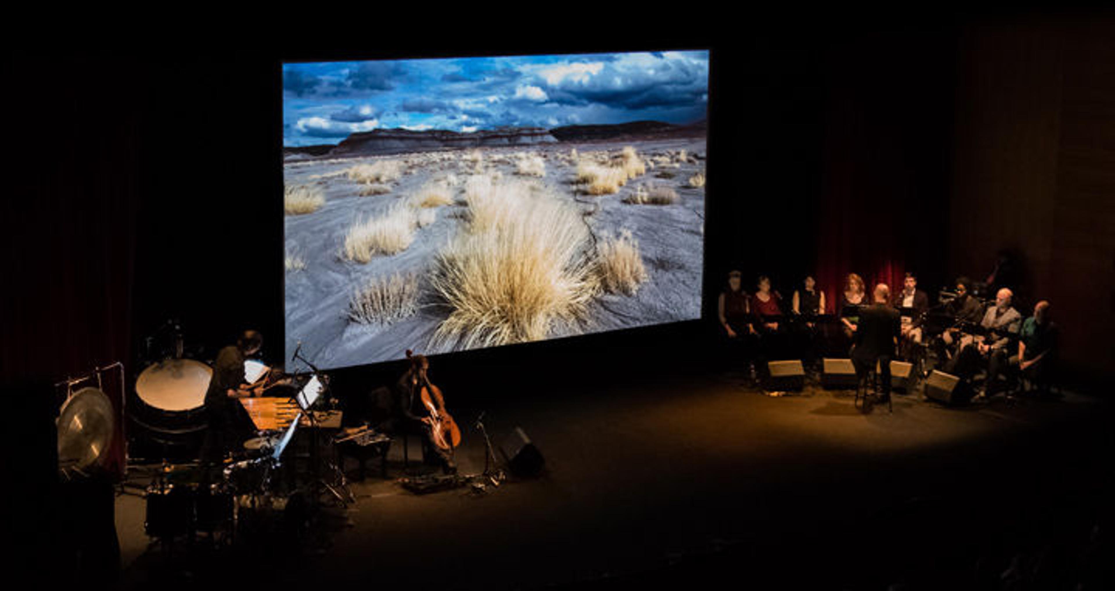 On a darkened stage several musicians and a chamber choir perform before a screen projection of a film depicting the Colorado River basin