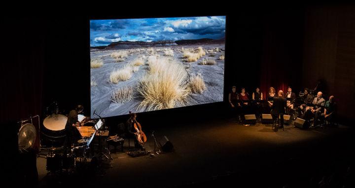 On a darkened stage several musicians and a chamber choir perform before a screen projection of a film depicting the Colorado River basin
