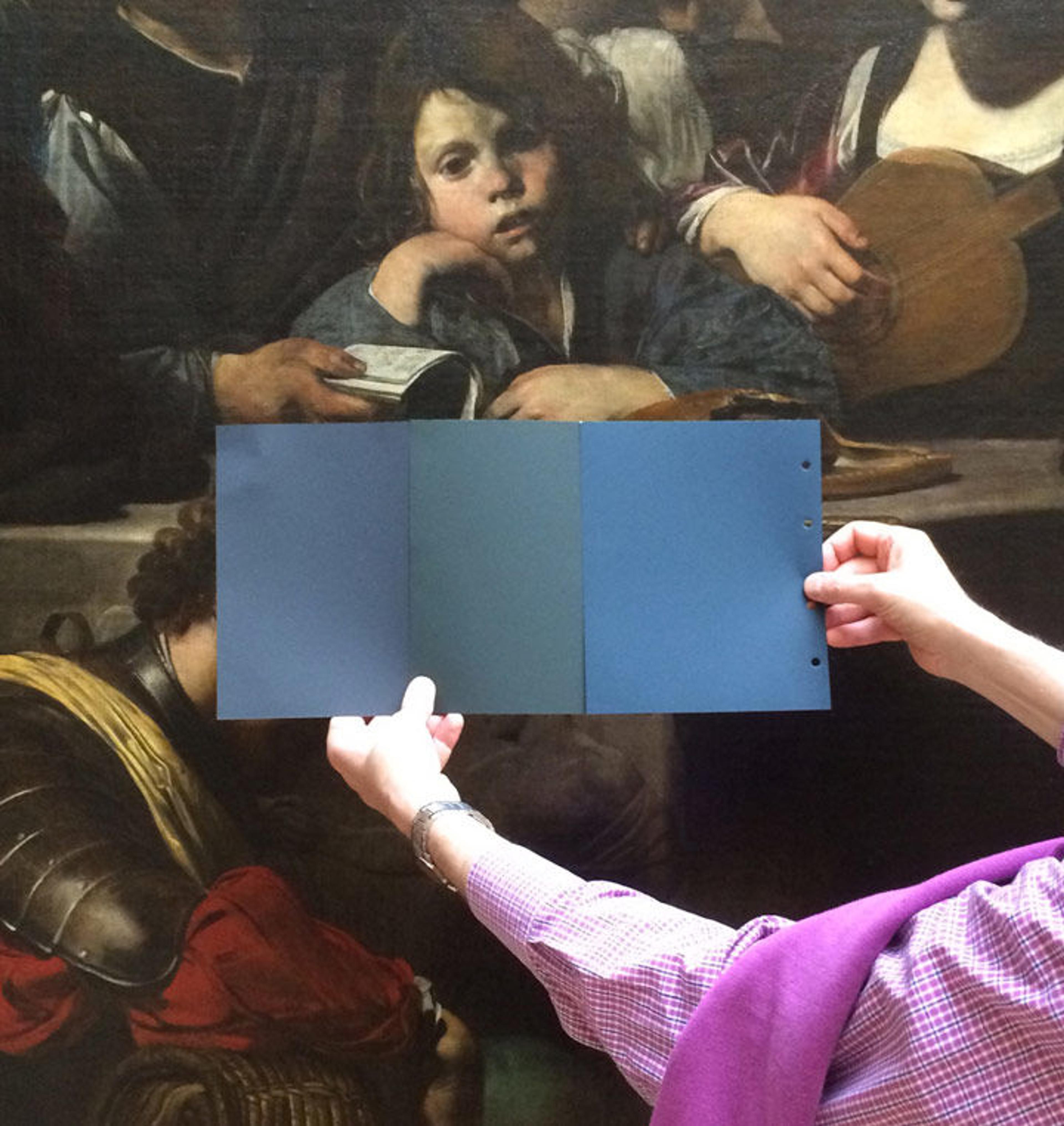Three swatches of blue are held near a painting of a domestic scene by the 17th-century artist Valentin de Boulogne