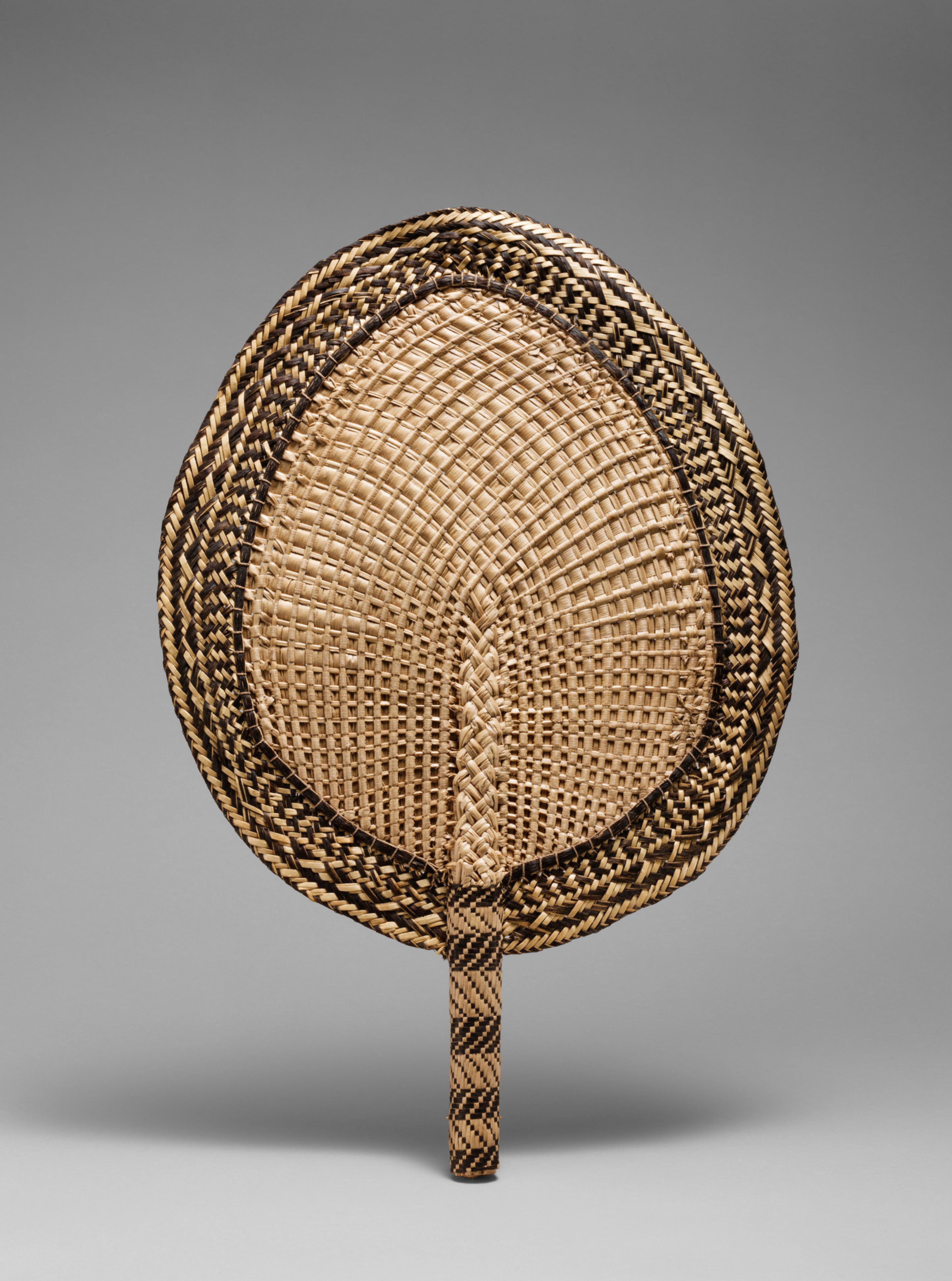 A woven handheld fan with a leaf-like shape and intricate black and natural straw patterns. The design is symmetrical, with a textured surface.