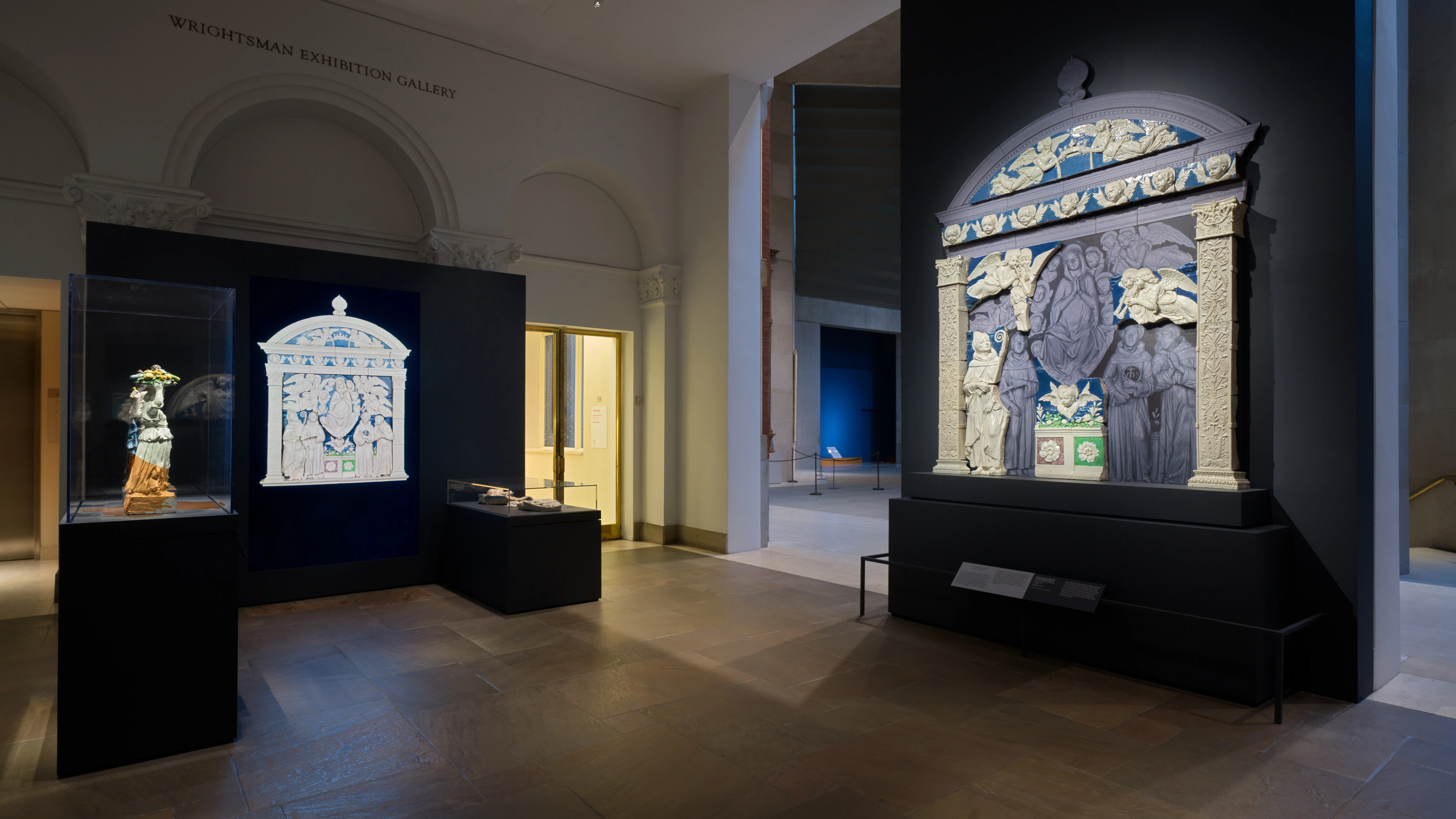 A quiet museum gallery displays an ornate blue-and-white arched panel with figures, alongside smaller illuminated artworks in dark cases.