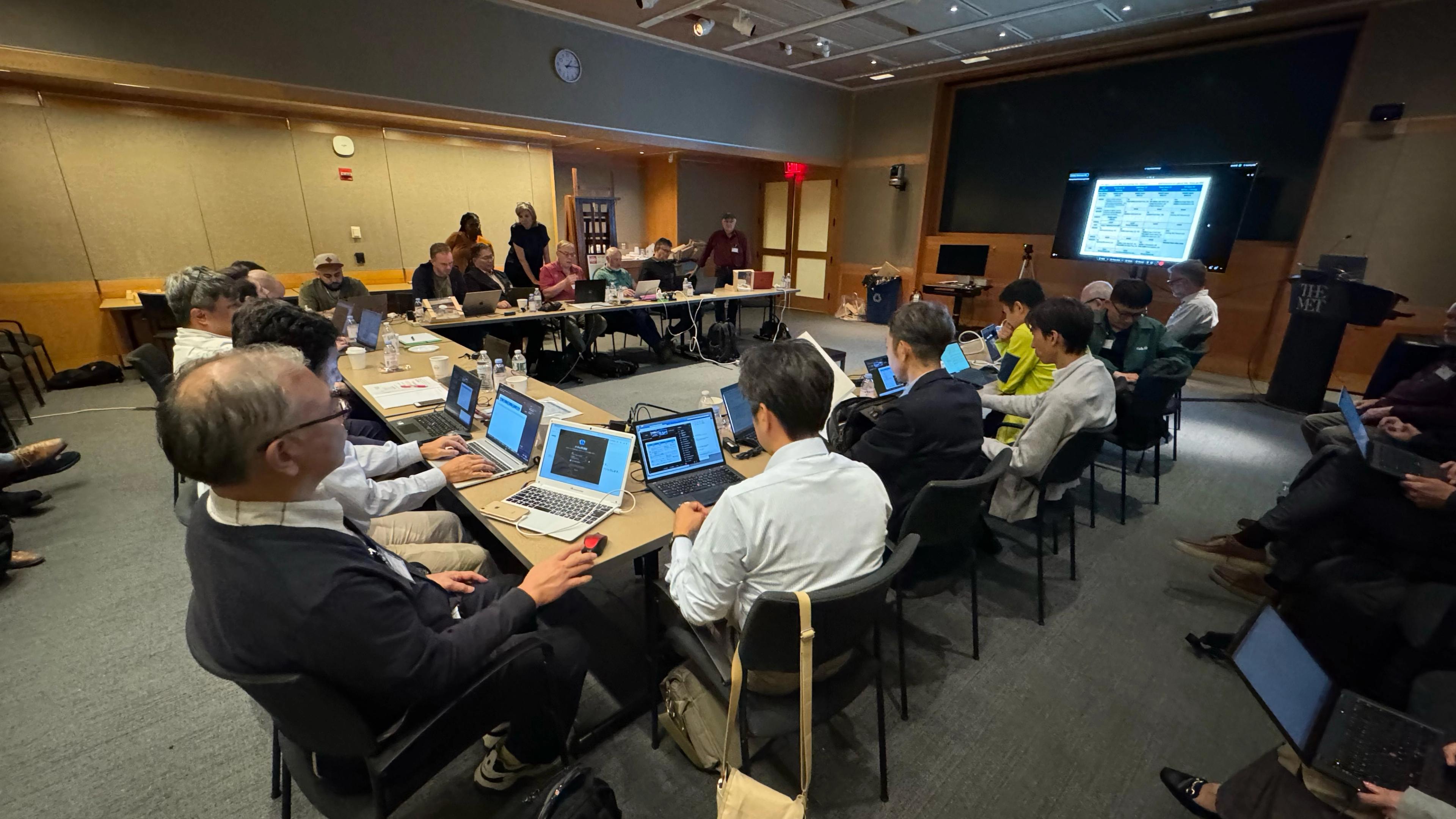 Numerous people with laptops sitting around a u-shaped table oriented towards a screen.