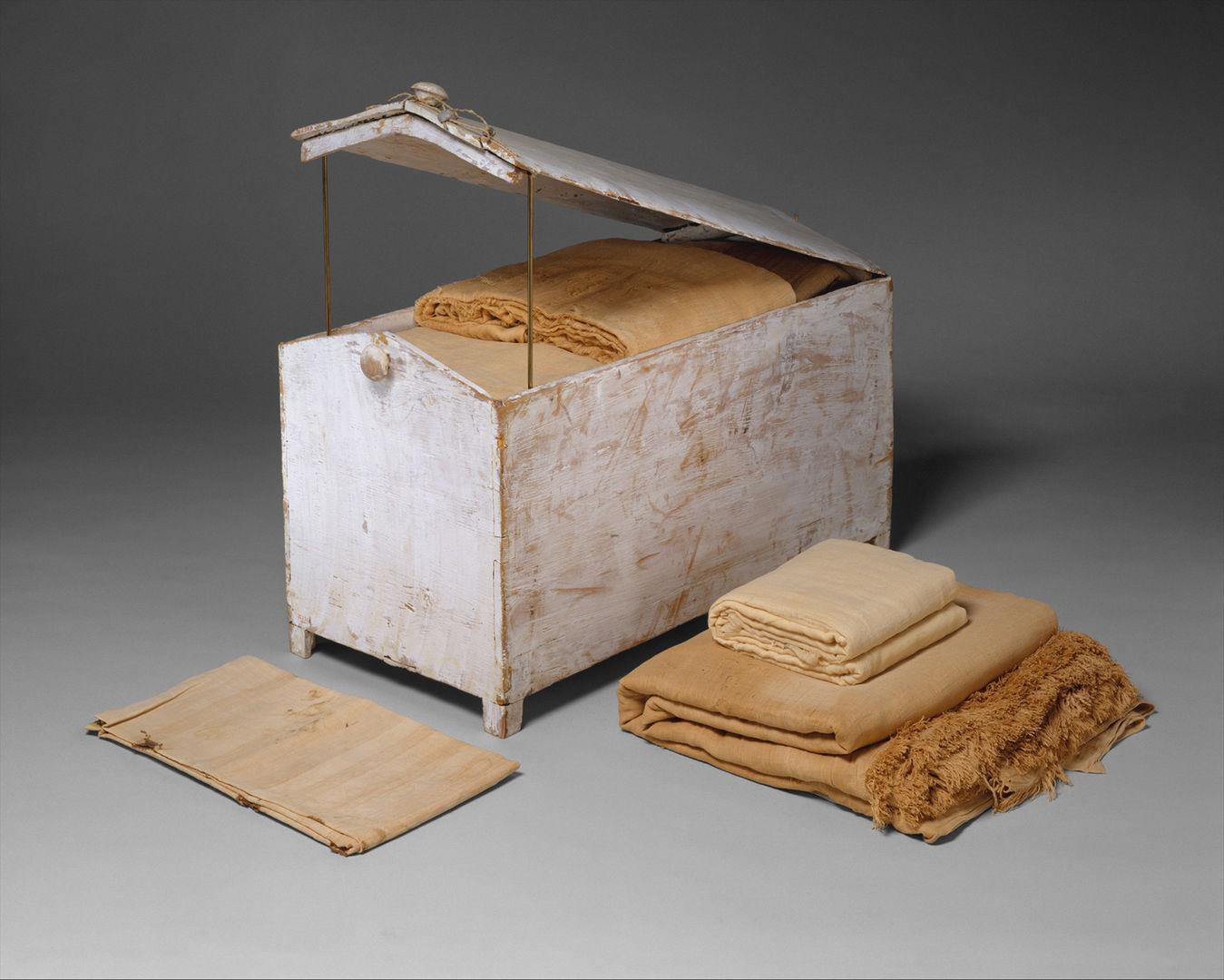 A gable-topped whitewashed chest with knobs for closure, shown partly opened to review folded linen inside. Several other folded cloths, all in various shades of beige, are around the chest.