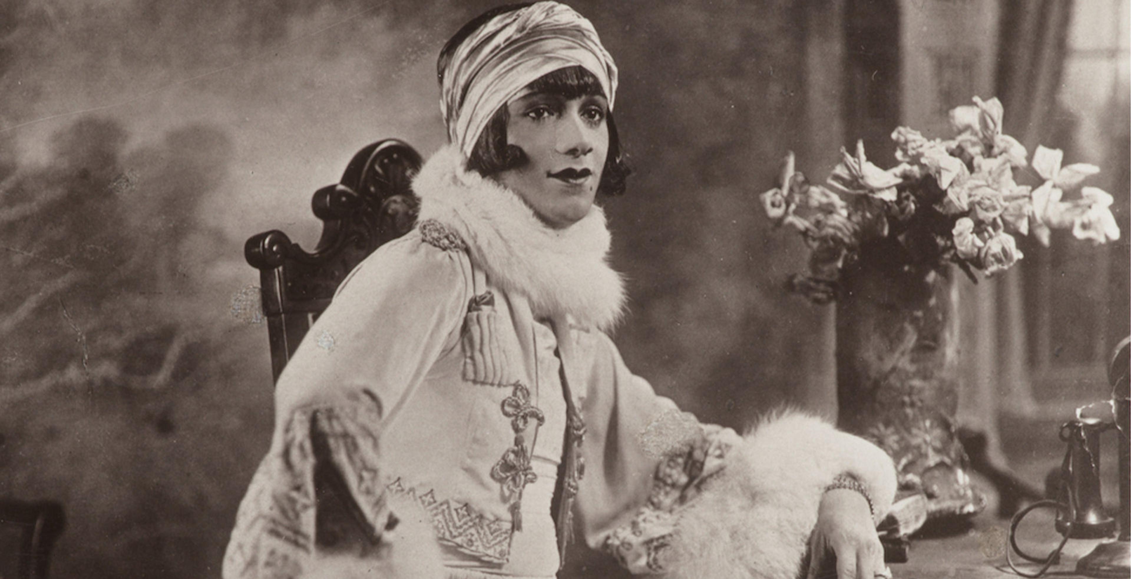 Woman sitting in wooden chair, facing the camera man. Calm expression and bold pose. 