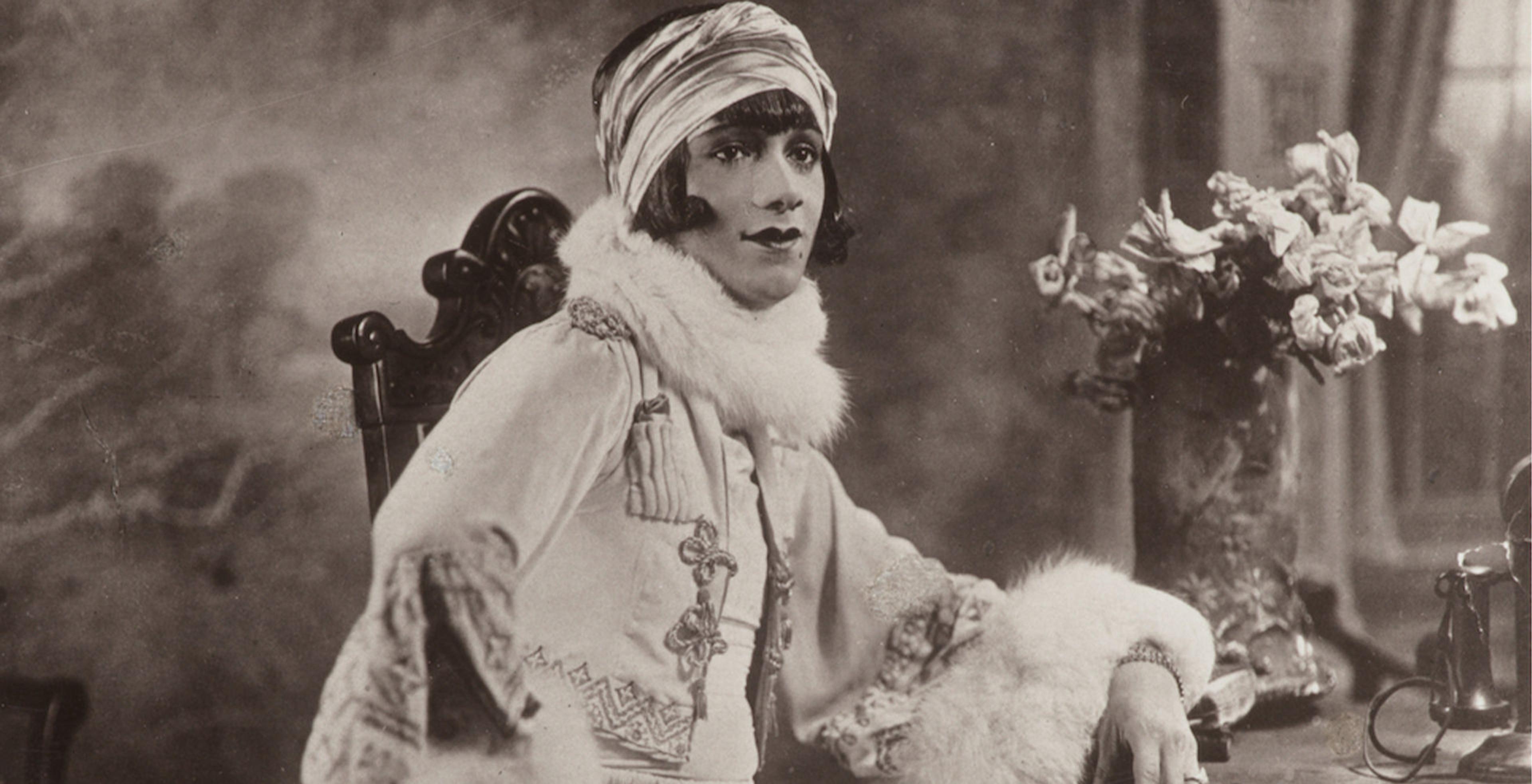 Woman sitting in wooden chair, facing the camera man. Calm expression and bold pose.