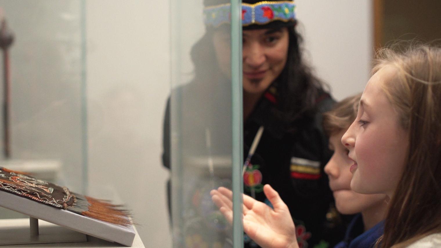 Artist Ty Defoe, Francesca, and Cyrus take a closer look at the Anishinaabe shoulder bag in the galleries. Francesca points out the detailed porcupine quill designs.