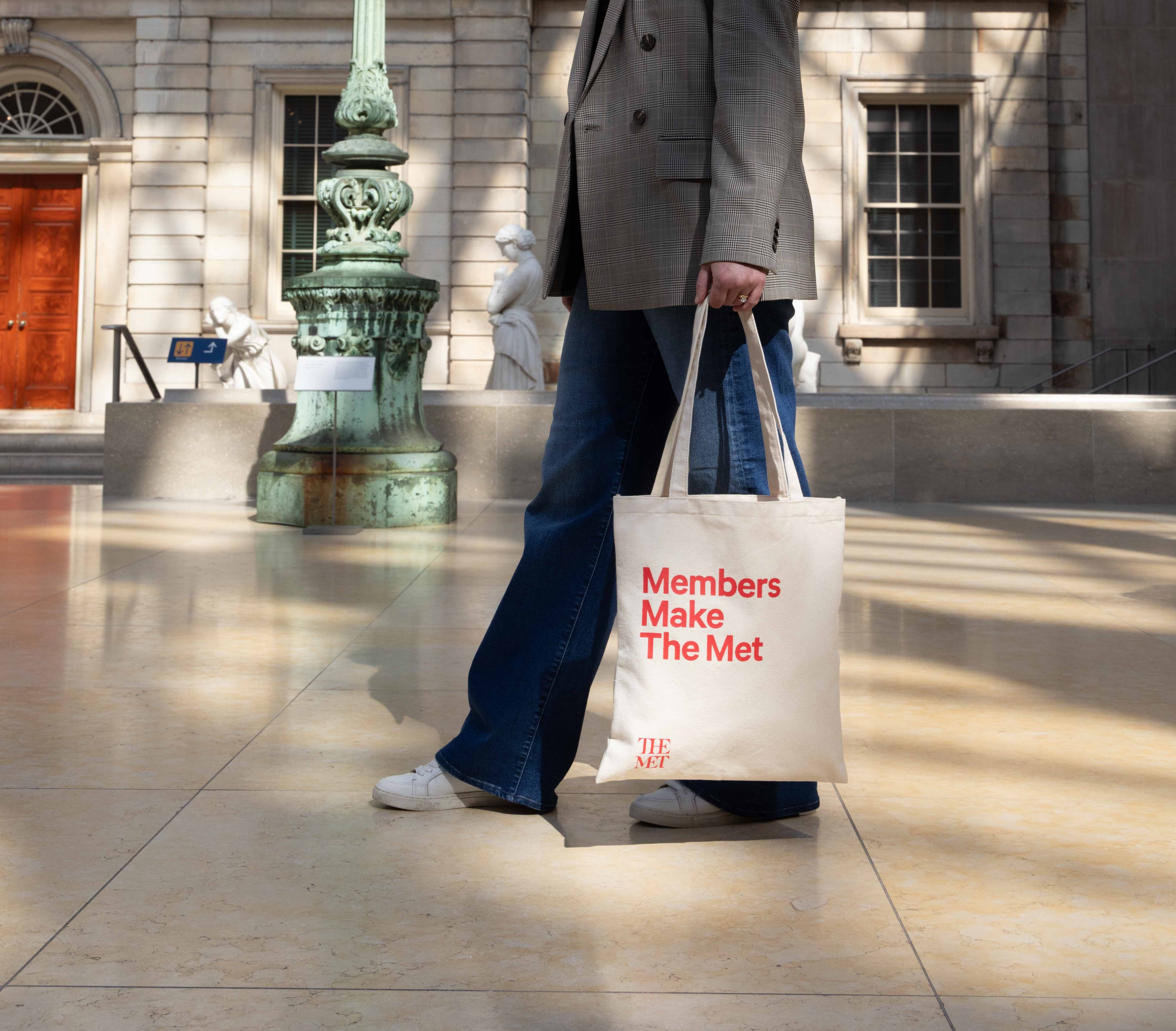 A cropped figure walks in The Met's American Wing with a Met Tote bag that reads "Members Make The Met"