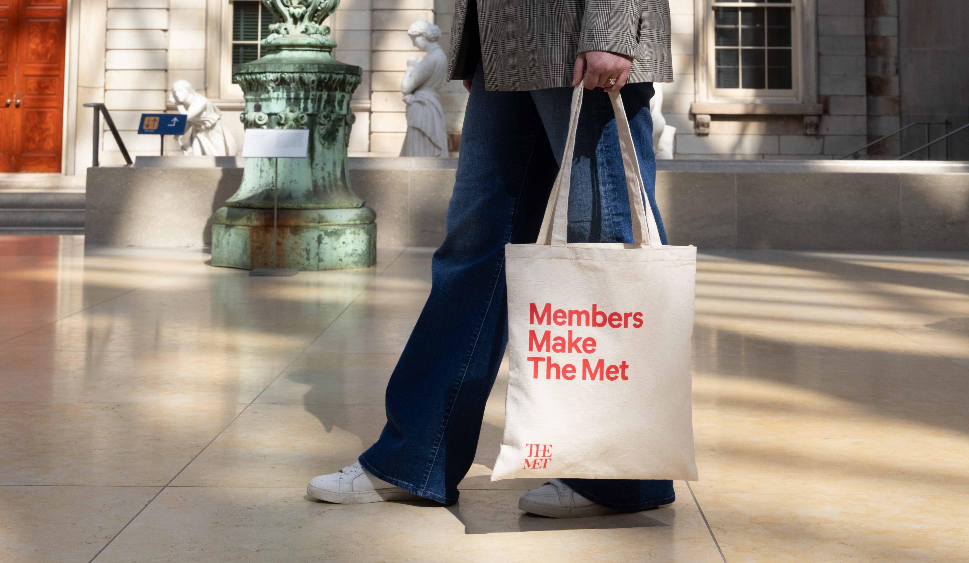 A cropped figure walks in The Met's American Wing with a Met Tote bag that reads "Members Make The Met"