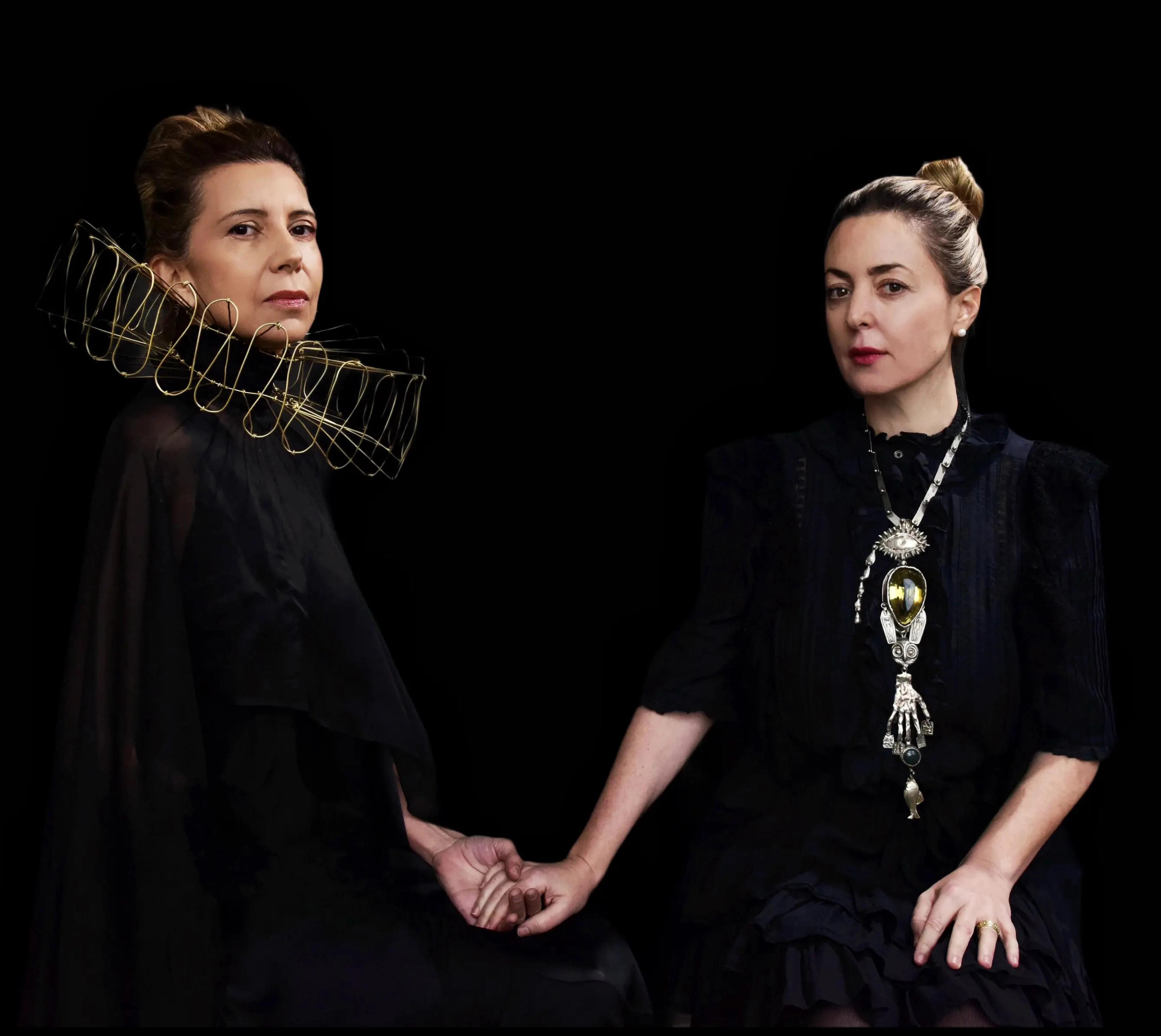 Paola Prestini and Magos Herrara, wearing black clothing dramatically accessorized with a collar and necklace, respectively, clasp each other's hand while staring at the viewer..