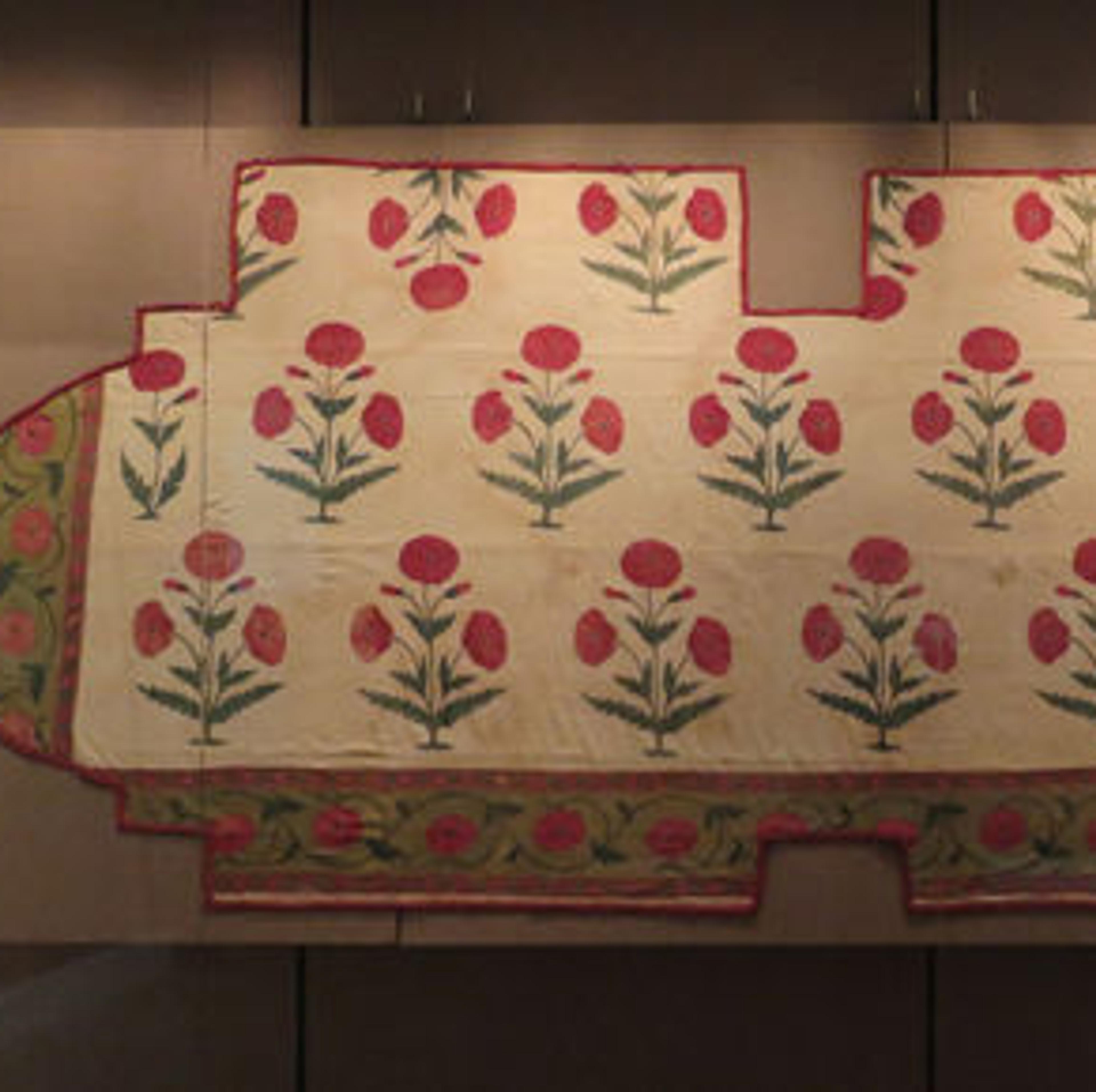 Large textile with a red flower pattern and green and red floral border, displayed flat. One small piece at right isn't attached to the rest of the fragment.