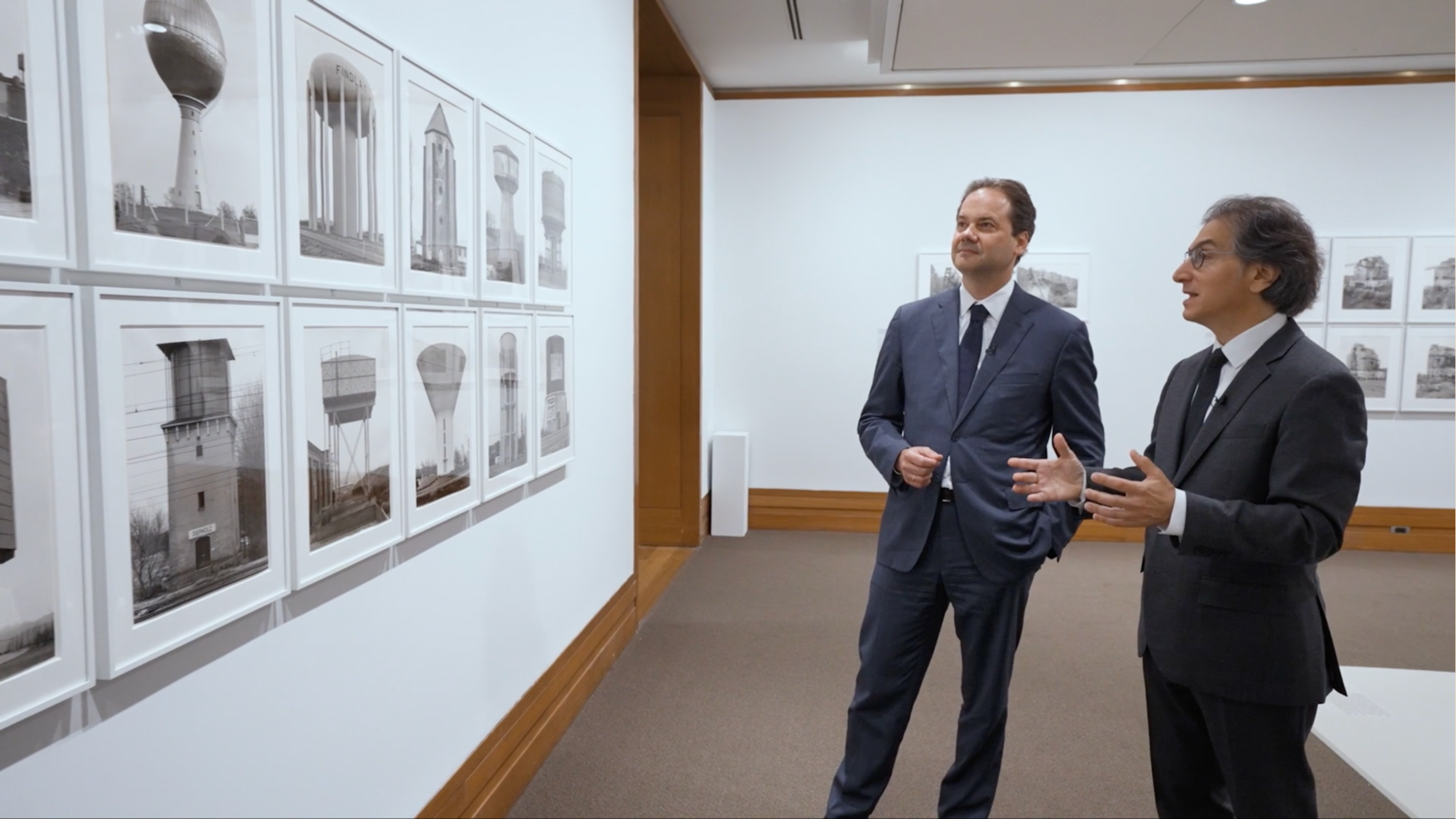 Two men in suits standing in gallery facing a wall of photographs.