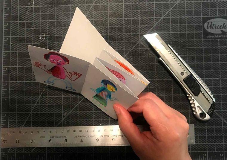 A hand pinches the folded paper over a cutting board. Beneath the hand is a steel ruler and above it is a craft knife.