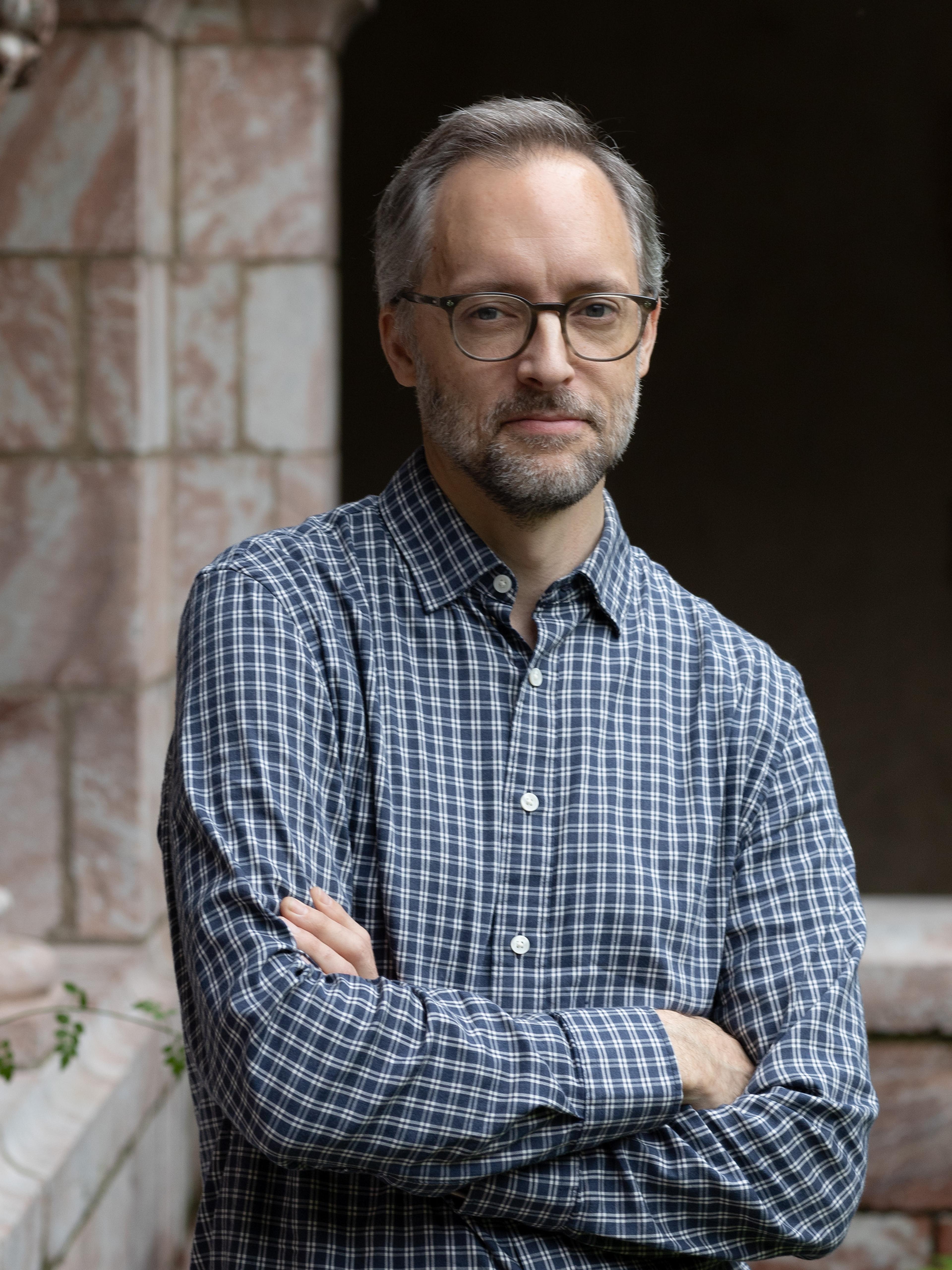 A man wearing a plaid shirt poses in front of a marble archway