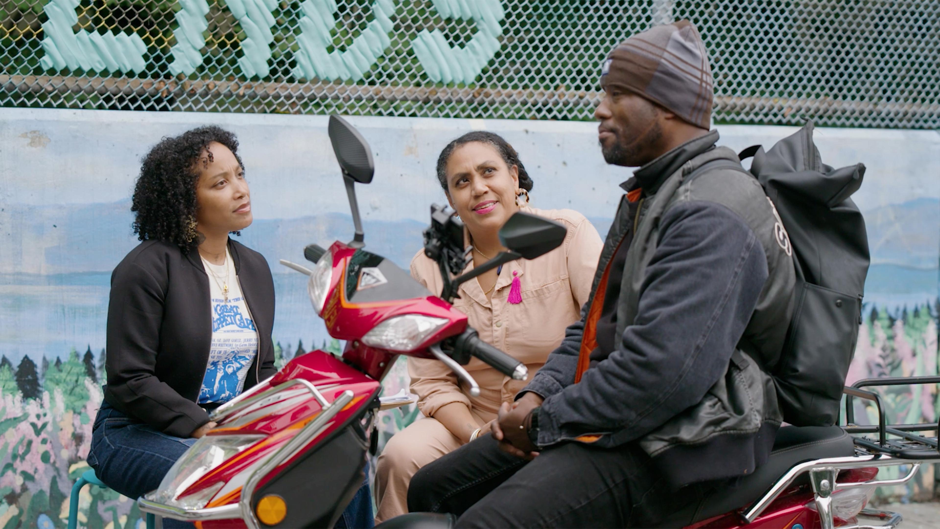 Mildred Beltré and Oasa DuVerney are seated outside, looking up at a man on a bike.