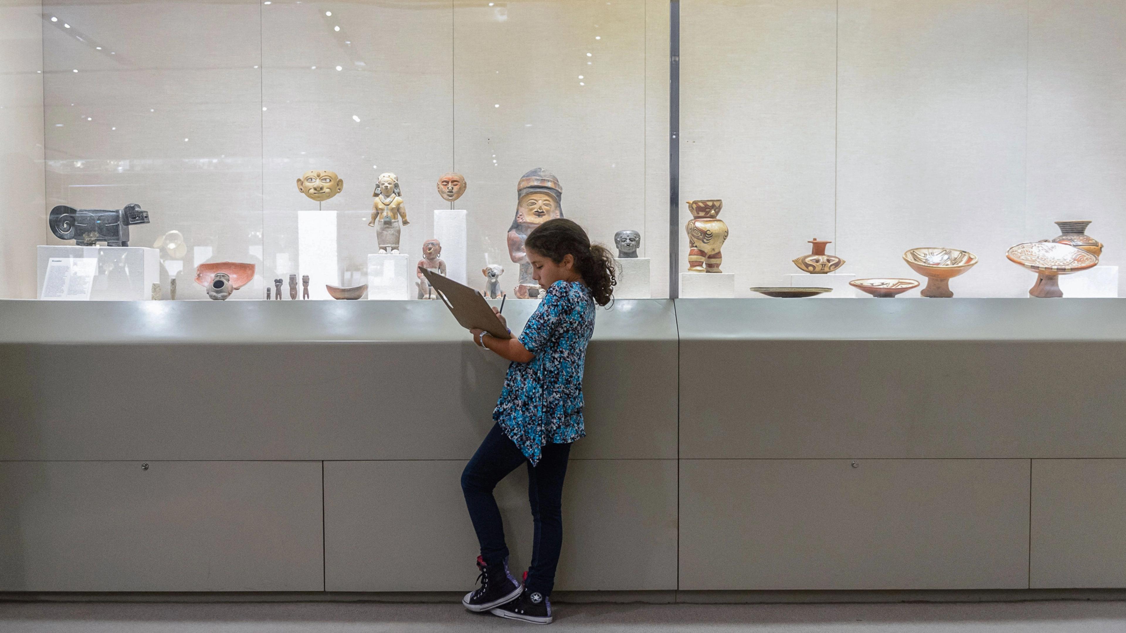 A young child holding a clipboard and pencil stands next to a glass case with ancient artifacts.
