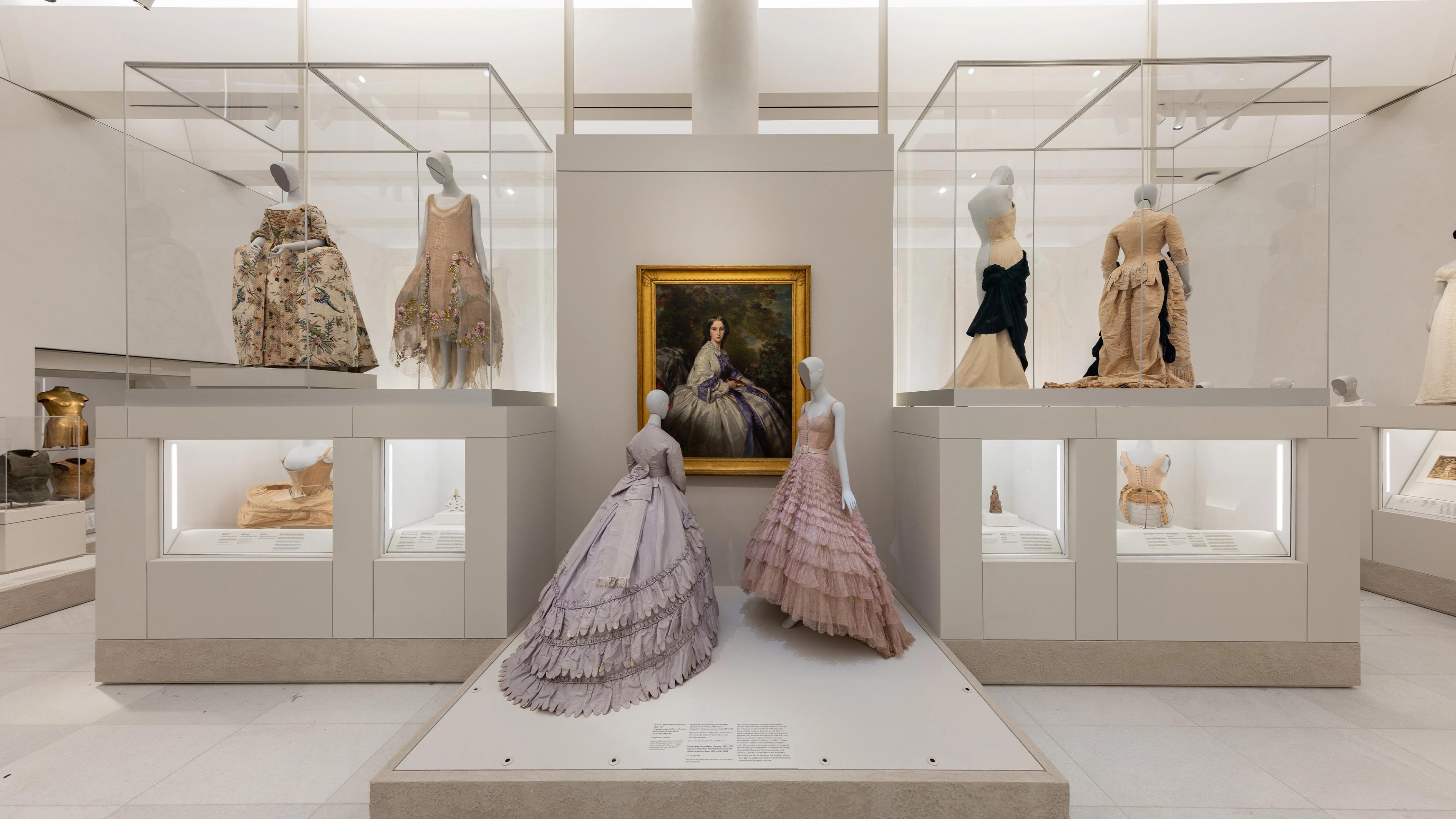 Exhibit of Victorian dresses on mannequins in a museum. Two pastel gowns in front of a portrait of a woman