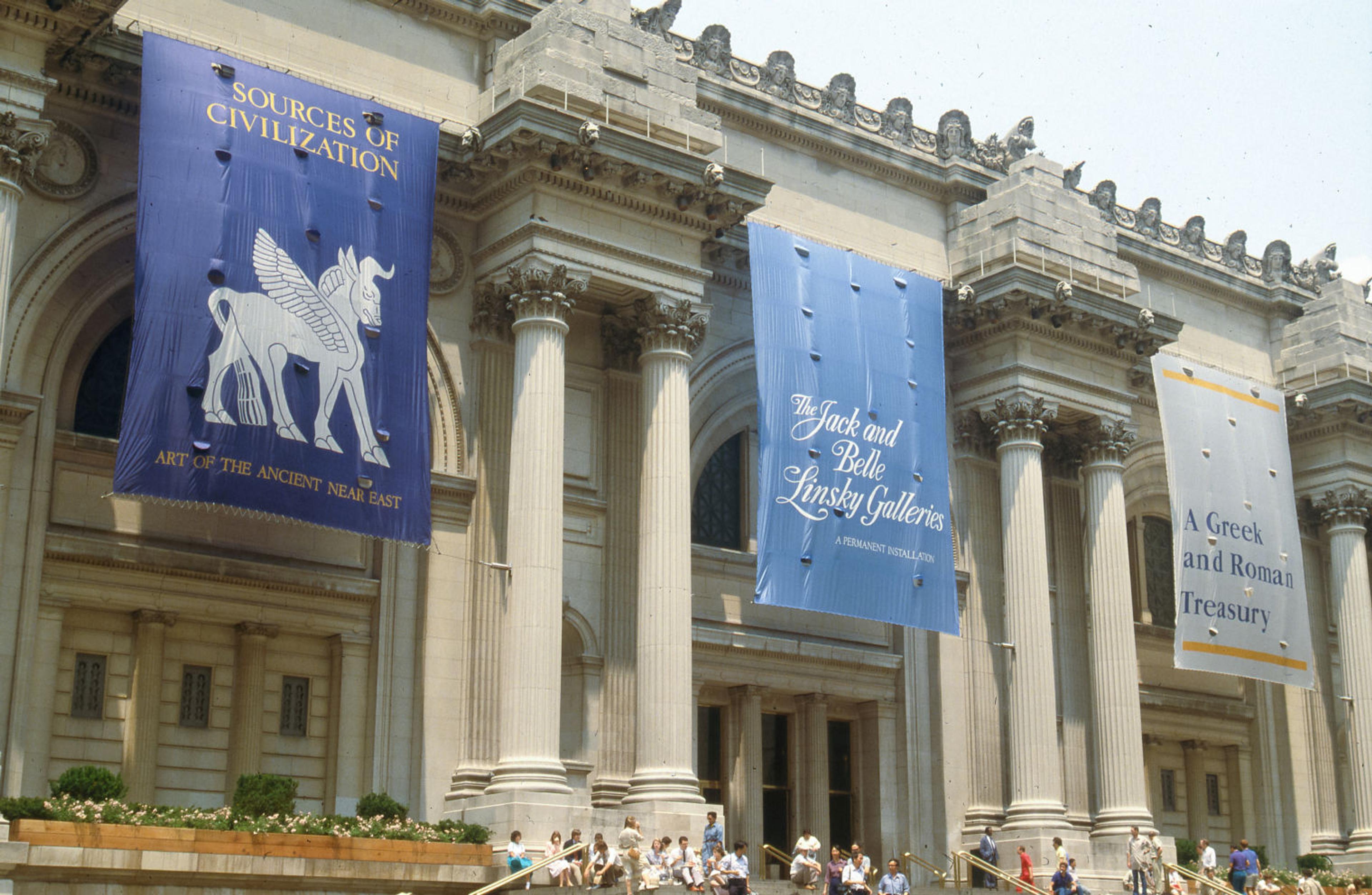 Entrance of the Metropolitan Museum of Art with people showing, left to right, banners for “The Ancient Near Eastern Art Gallery,” “The Jack and Belle Linsky Collection,” “Greek and Roman Art Gallery”