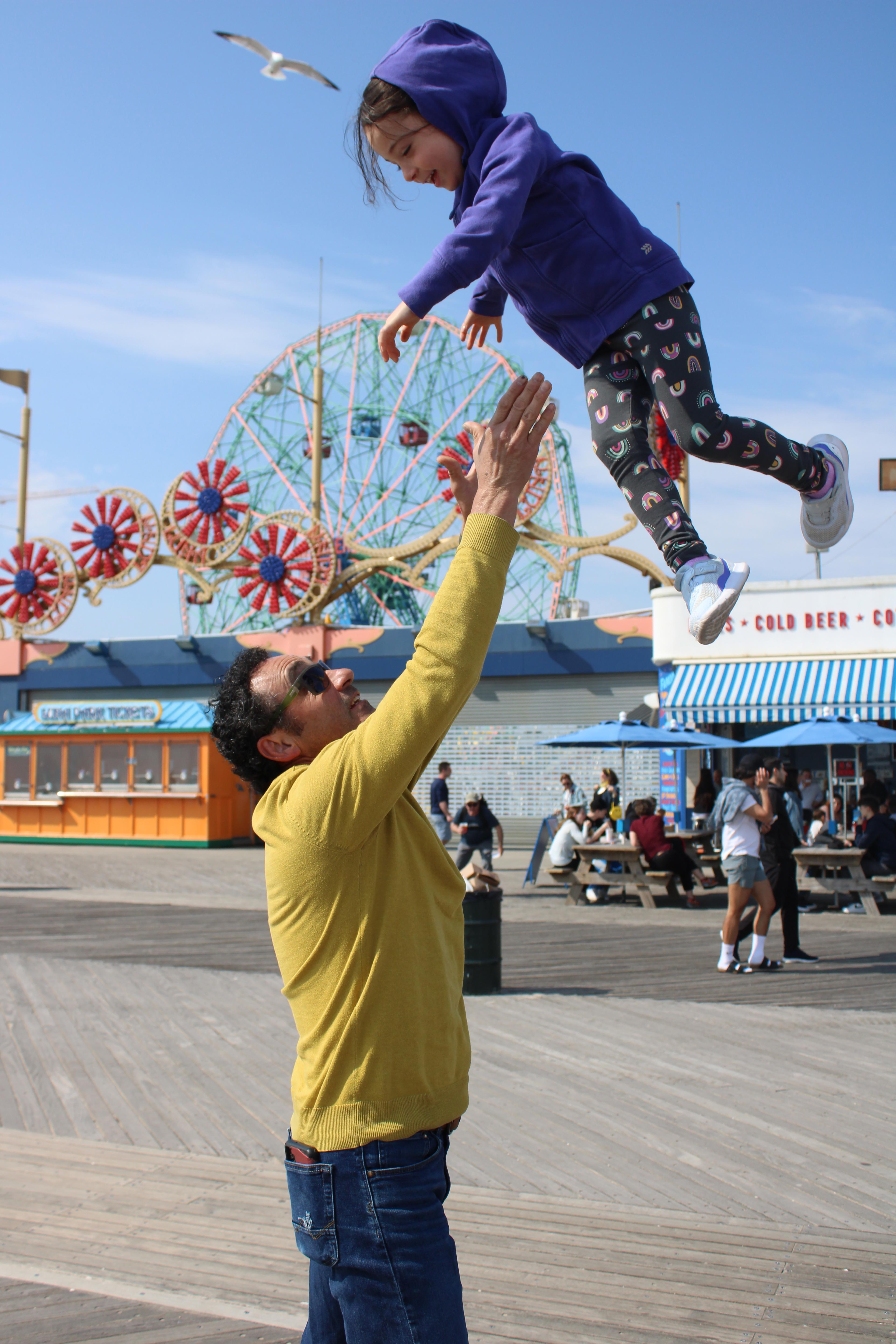 Color photograph of a man wearing a yellow sweatshirt, sunglasses, and blue jeans hoisting a small girl aloft with both hands in the air above him on the Coney Island boardwalk during the day. There are concession stands and a Ferris wheel in the background. The girl is smiling and gazing down at the man below. She wears a blue hoodie jacket, black leggings covered in decorative patterns, and white sneakers. The sky is blue and a seagull flies overhead in the distance, to the left of the girl's head.