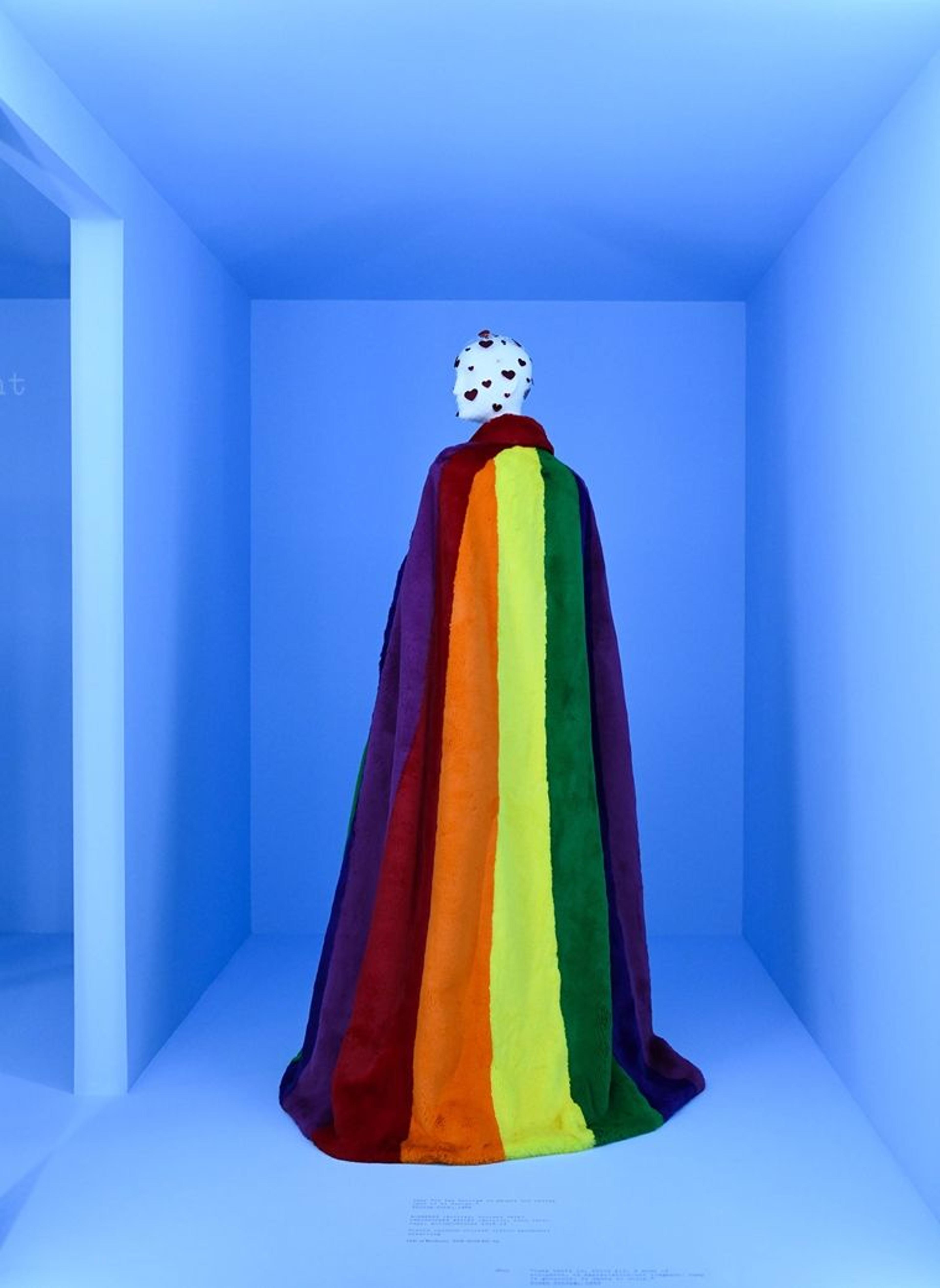 An installation view of a mannequin wearing a fuzzy Burberry cape in a bright rainbow design.