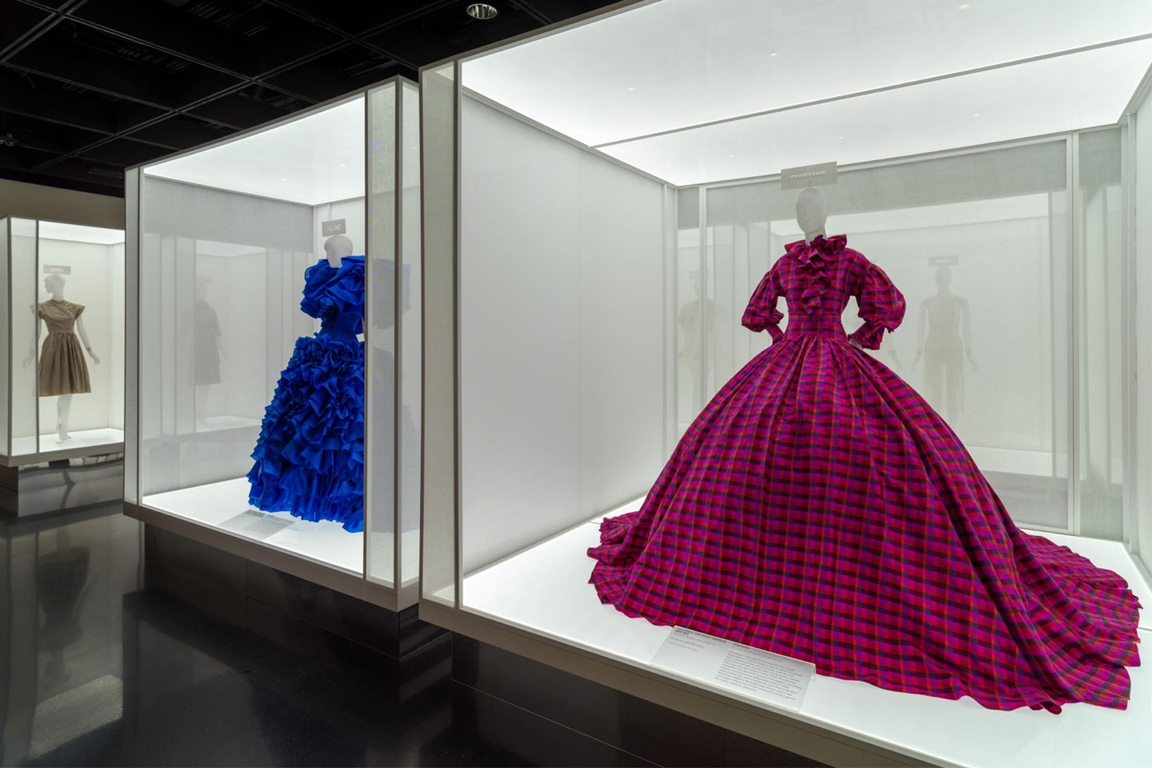 An in-gallery view of dresses presented in illuminated light box cases; the dress appearing closest in the foreground has a bight fuchsia tartan pattern with long sleeves, fitted bodice and large crinoline skirt.
