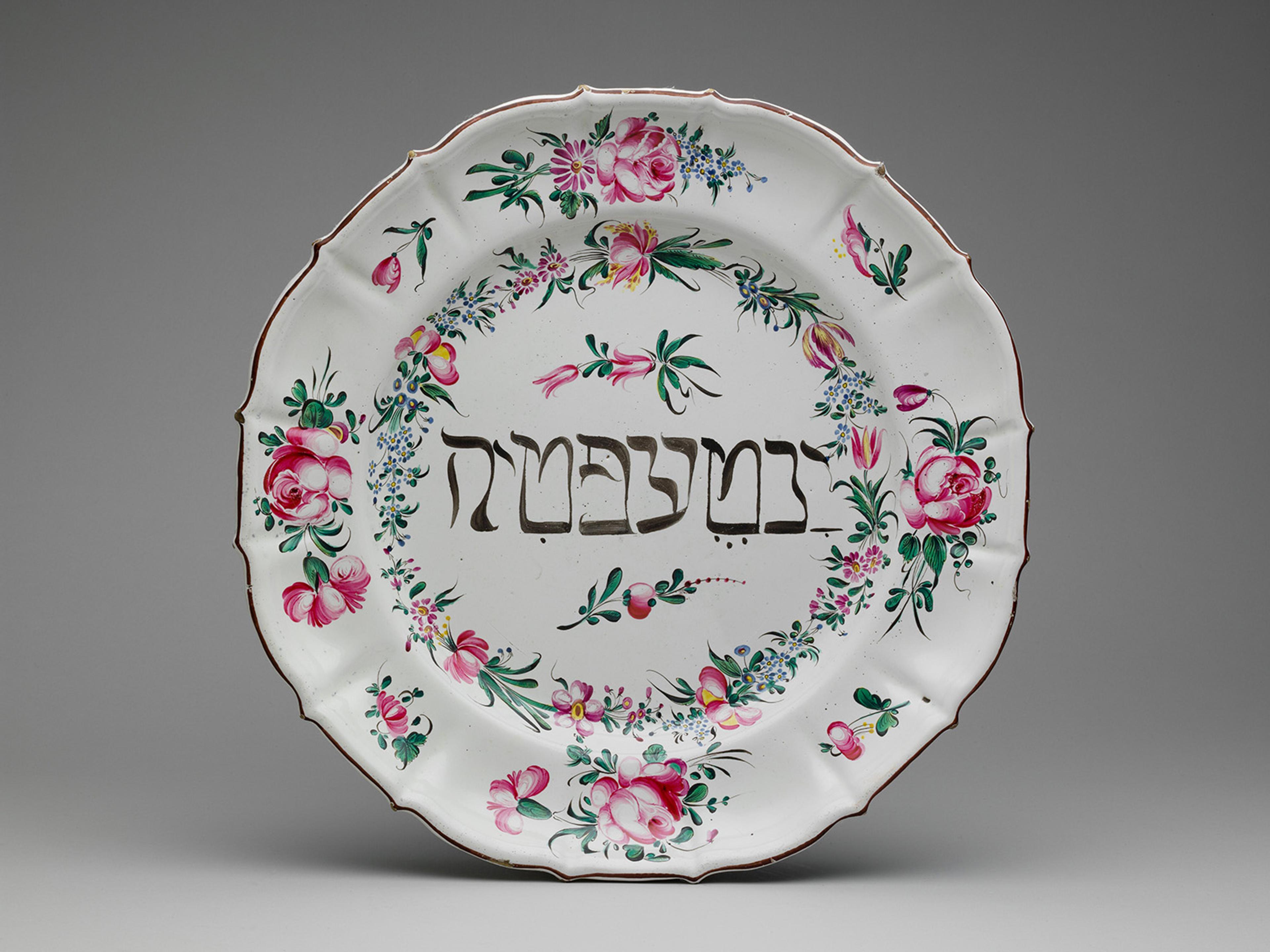 Ornate plate with Hebrew script at center, surrounded by vibrant pink floral patterns and green leaves on a white background.