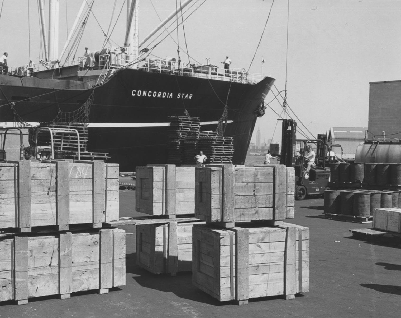 Black and white photograph of wooden crates stacked on a pier with the ship Concordia Star in the background