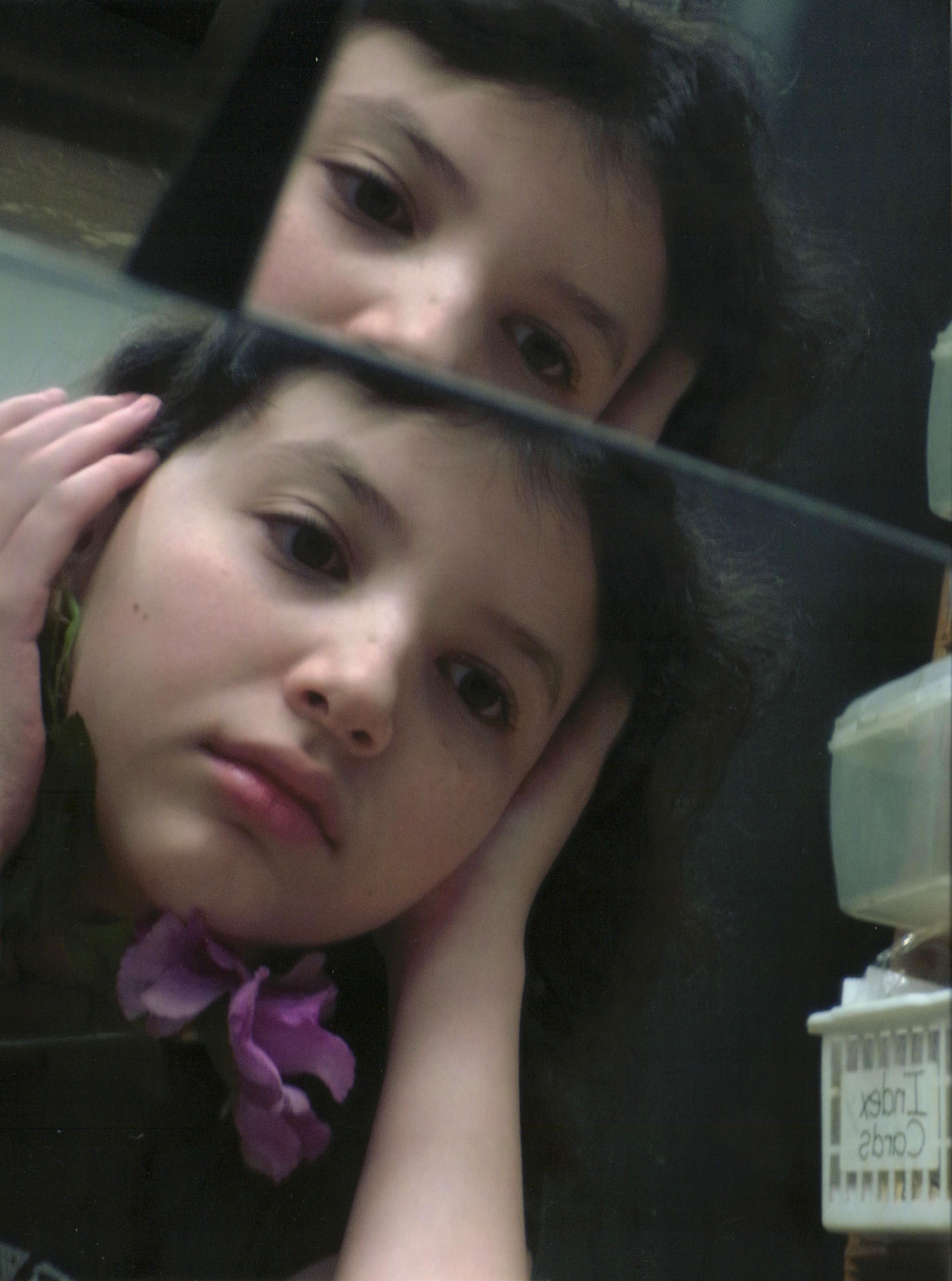Color close-up photo of a young white girl with dark hair, captured in the reflective surfaces of two mirrors, one directly in front of the other. In the lower mirror, the girl is holding both hands over her ears and facing right, away from the camera. A violet flower dangles horizontally just beneath her chin. In the top mirror, only the nose, eyes, and hair on top of the girl are visible. A small, white plastic container is cropped out on the lower right side of the bottom mirror. The words "Index Cards" appear reversed in the mirror's reflection on a small sign affixed to the container.