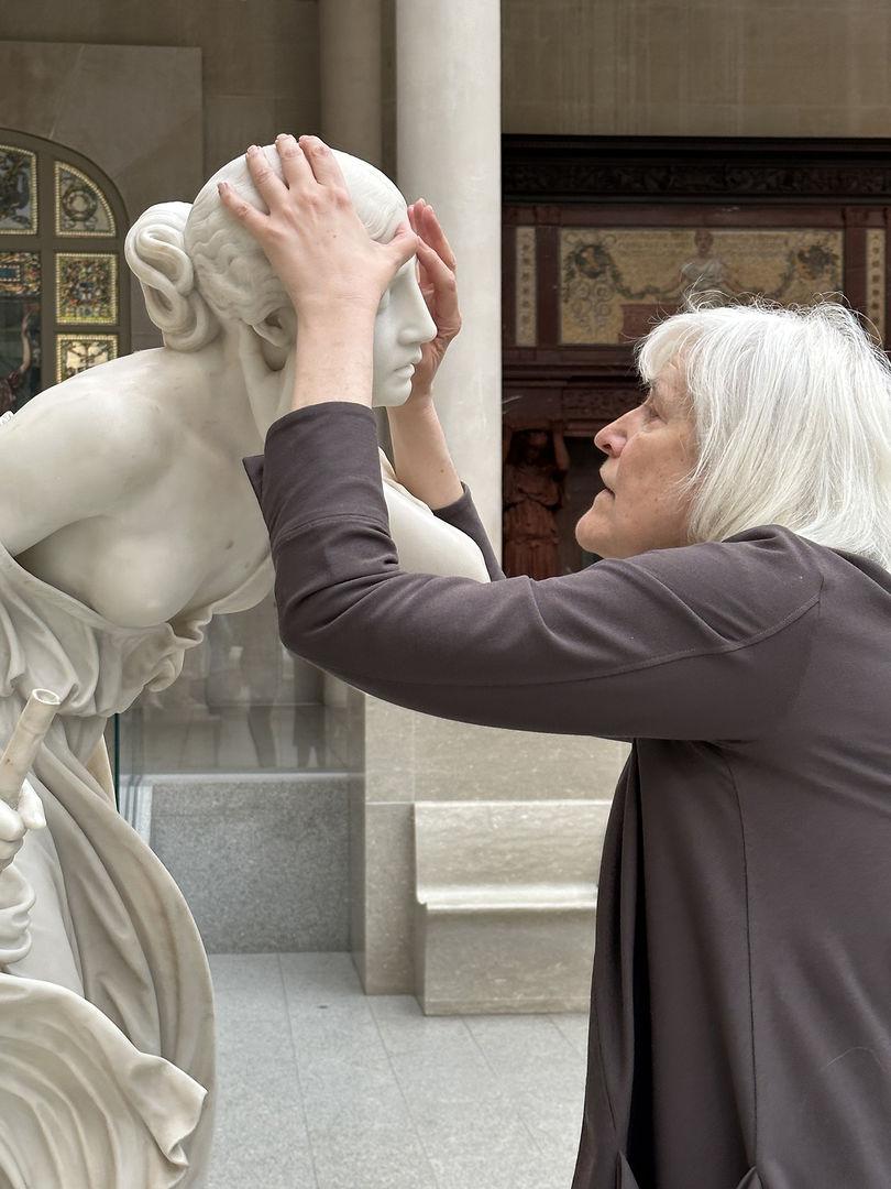 Image of the writer and professor Georgina Kleege, with her hands around the head of Nydia, The Blind Flower Girl of Pompeii, during a touch tour in the American Wing Engelhard Sculpture Court at The Met, a skylit space with direct, dramatic natural light. The image depicts the profile of a grey-white-haired woman with medium long straight hair with a long-sleeved brownish purple sweater touching the face of an almost life-sized white marble statue of a young girl. Her left hand is on the top of the sculpture's head, and her right thumb is touching the forehead. Two decorative artworks are installed into the wall in the background: one stained glass window and a fireplace.