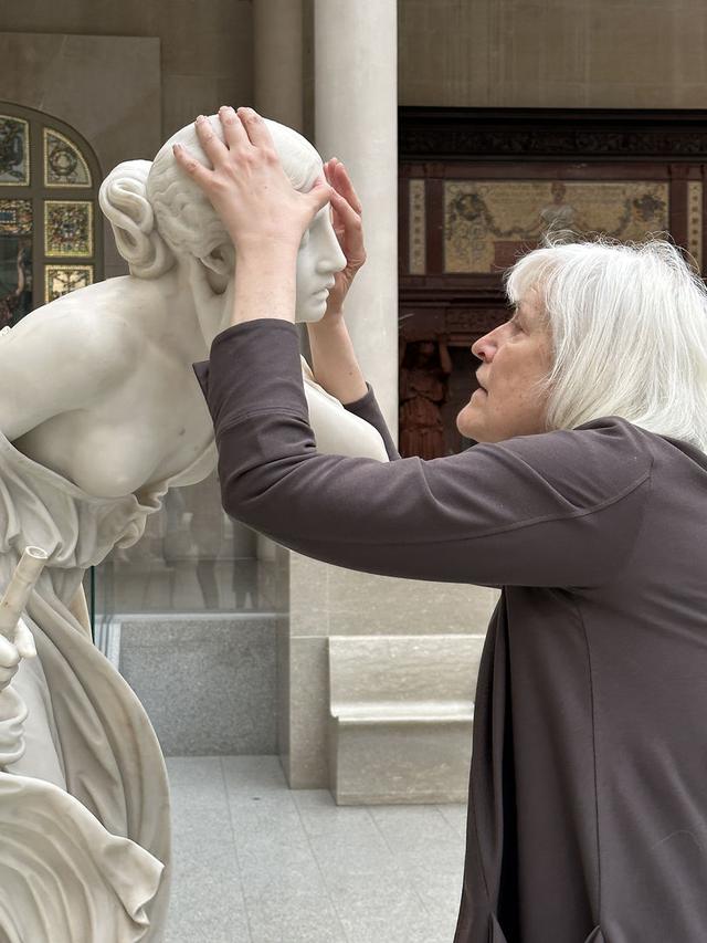 Image of the writer and professor Georgina Kleege, with her hands around the head of Nydia, The Blind Flower Girl of Pompeii, during a touch tour in the American Wing Engelhard Sculpture Court at The Met, a skylit space with direct, dramatic natural light. The image depicts the profile of a grey-white-haired woman with medium long straight hair with a long-sleeved brownish purple sweater touching the face of an almost life-sized white marble statue of a young girl. Her left hand is on the top of the sculpture's head, and her right thumb is touching the forehead. Two decorative artworks are installed into the wall in the background: one stained glass window and a fireplace.