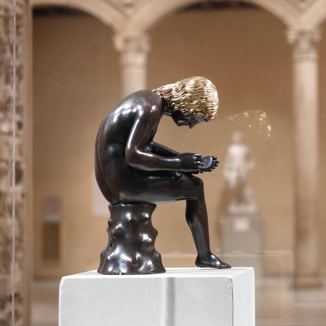 A small bronze sculpture of a seated boy pulling a thorn from the bottom of his foot appears within a display case at The Metropolitan Museum of Art; the figure hunches over looking intently at the bottom of his foot; the sculpture is set against the warm walls and limestone columns of the against gallery in the distance.