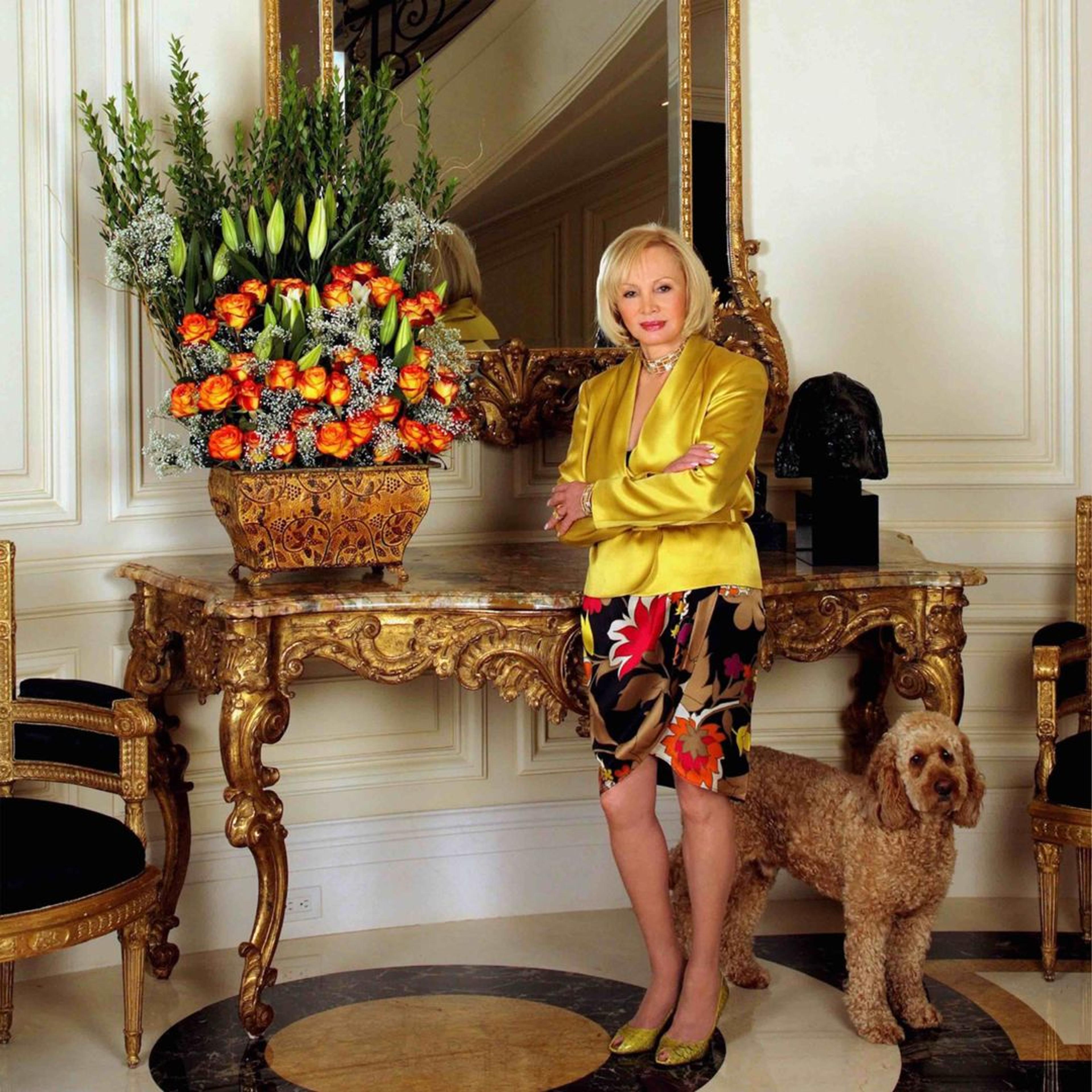 A woman with short blonder hair and a yellow vest stands with a dog to her left in front of an armoire