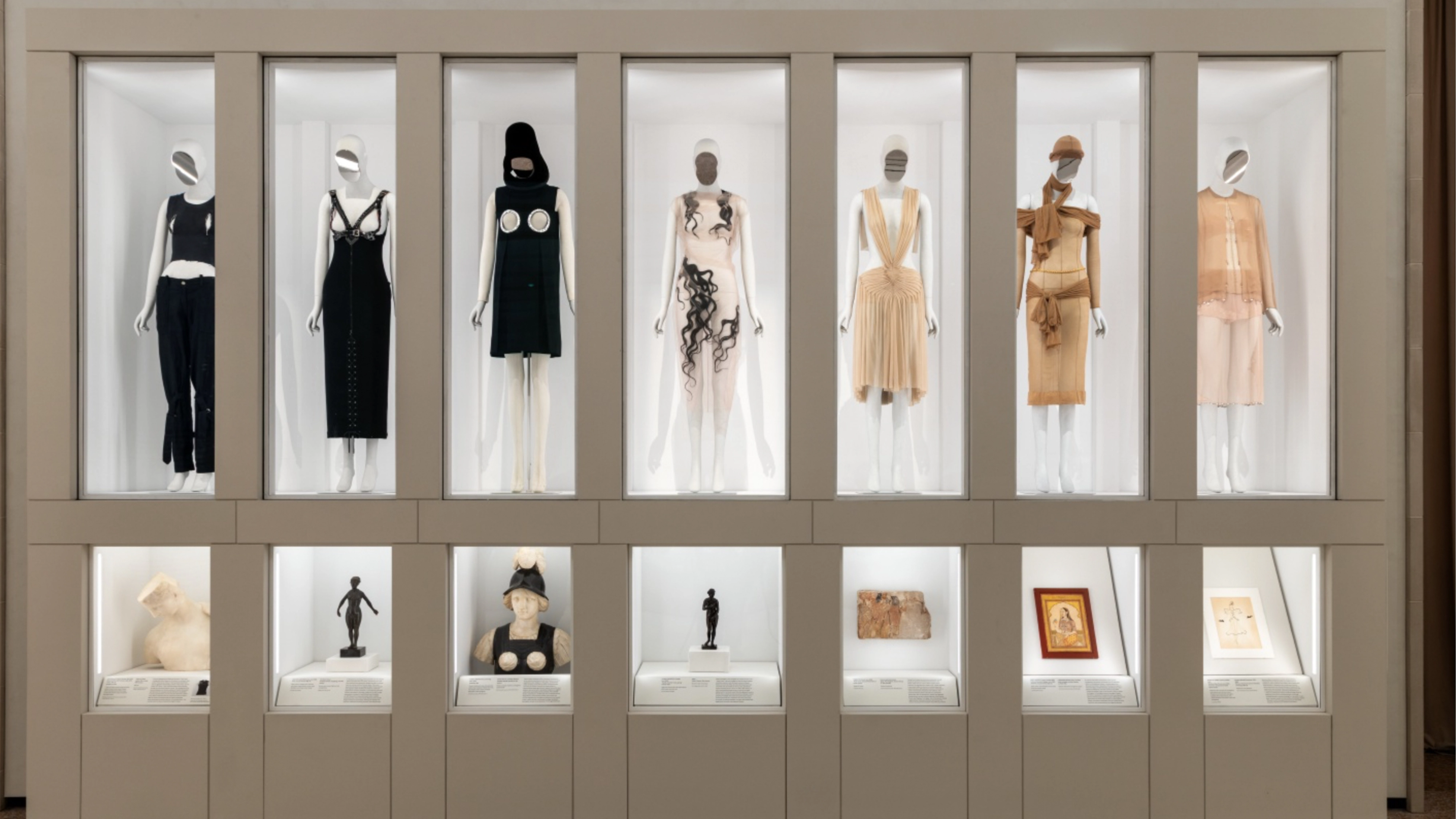 A museum display presents historic dresses and outfits on mannequins above glass cases of fashion artifacts.