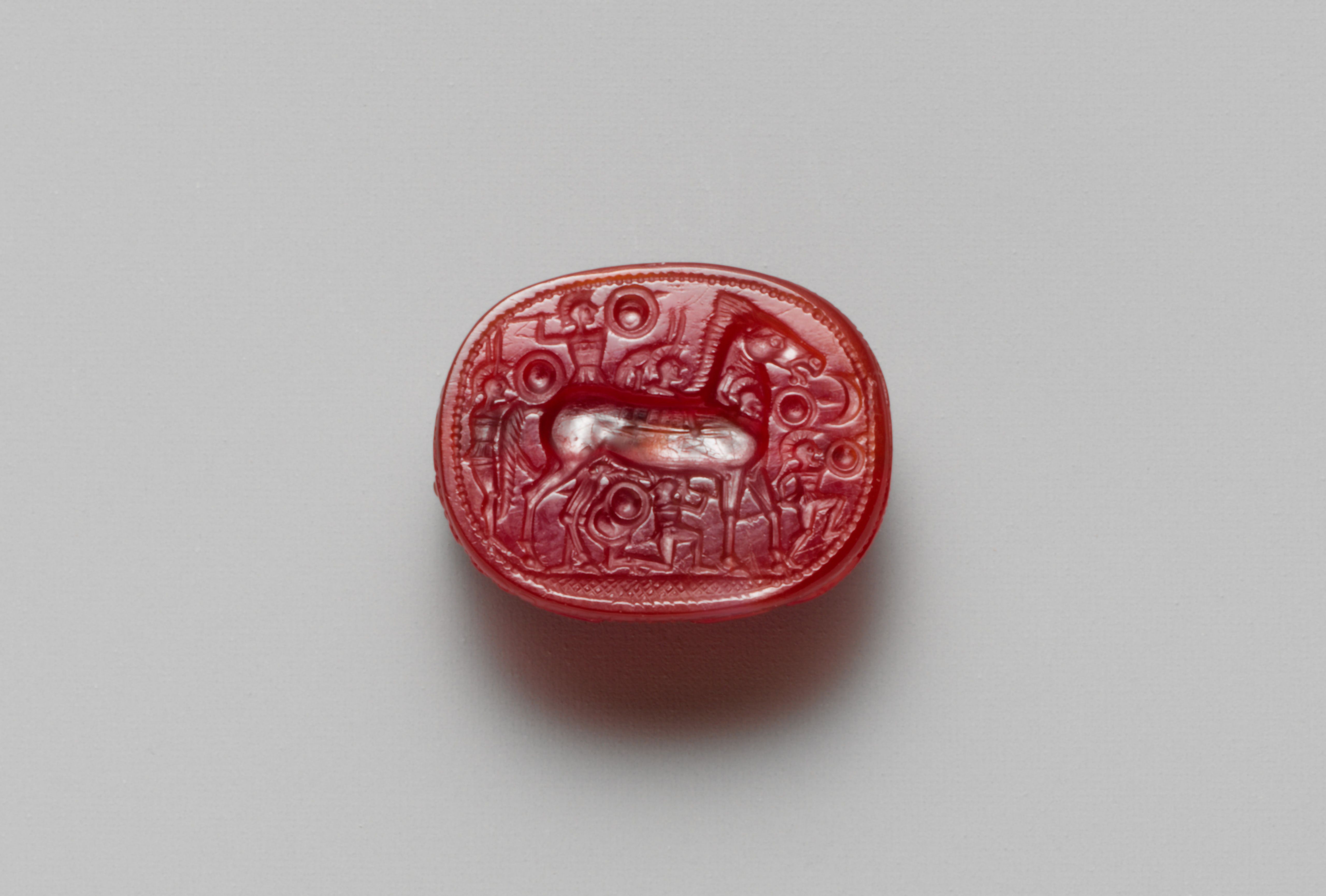 Small red stone, intricately carved with a horse and shield-wielding fighters.