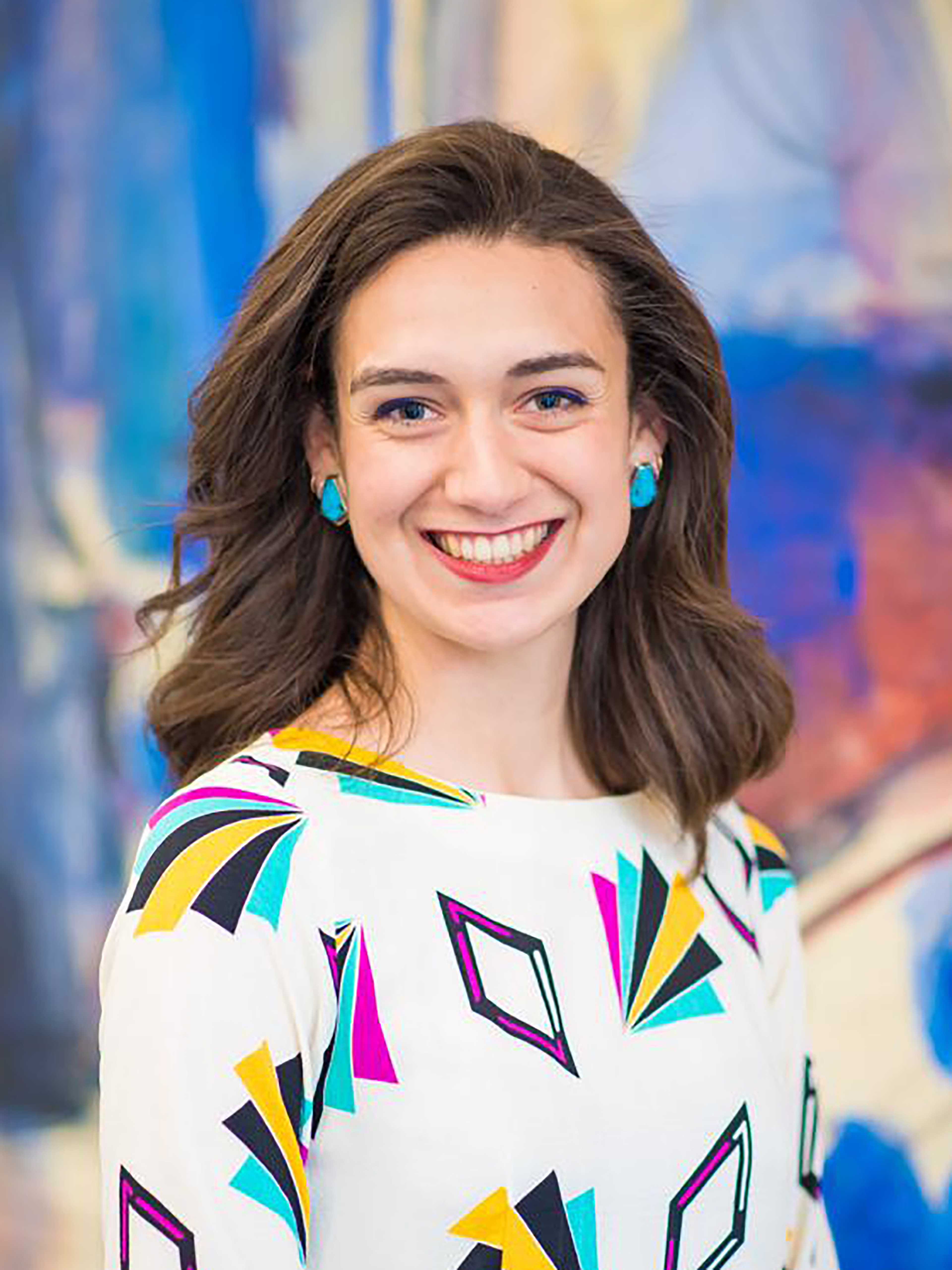 A woman wearing a colorful shirt and earrings poses in front of a blue painting