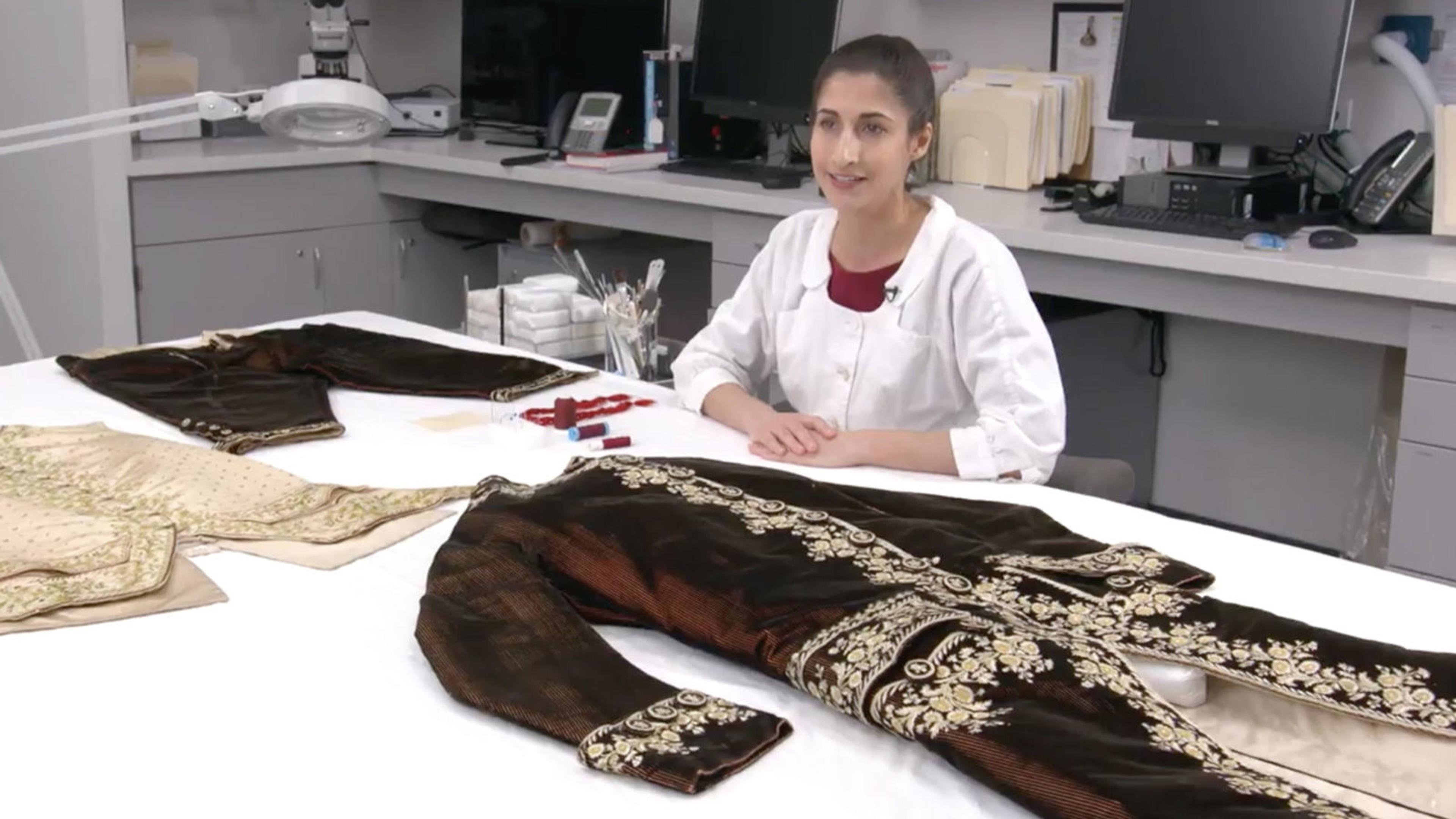 A conservationist sits at a white table. Laid out on the table in front of her are different pieces of an 18th century men's suit.