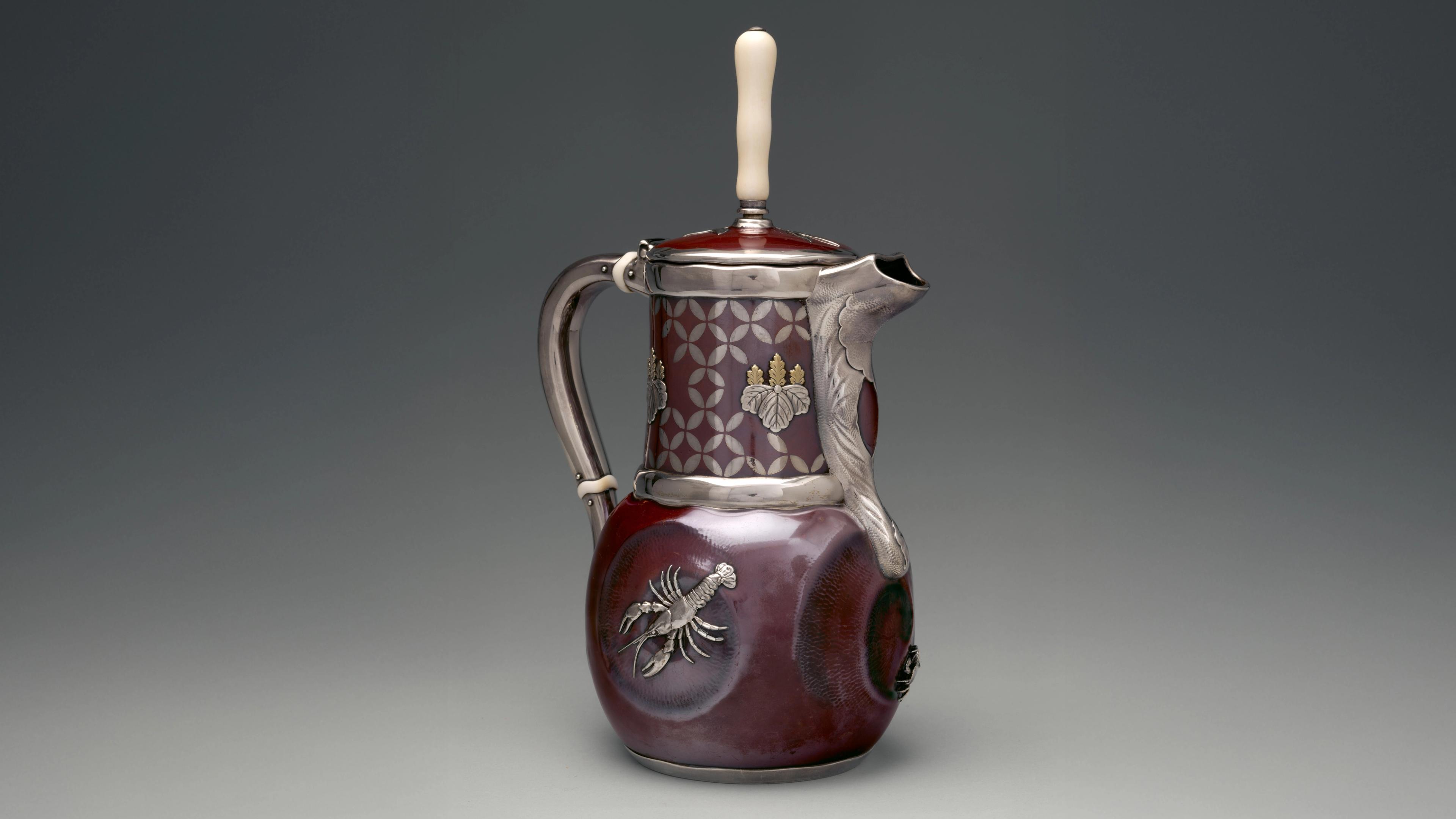 A three-quarter view of a patinated copper and silver chocolate pot in the shape of a pitcher with a handle, spot, and ivory handle. Silver banding emphasizes the handle, spout, and rim of the lid and neck. A lobster rendered in high-relief silver adorns the body of the pot.