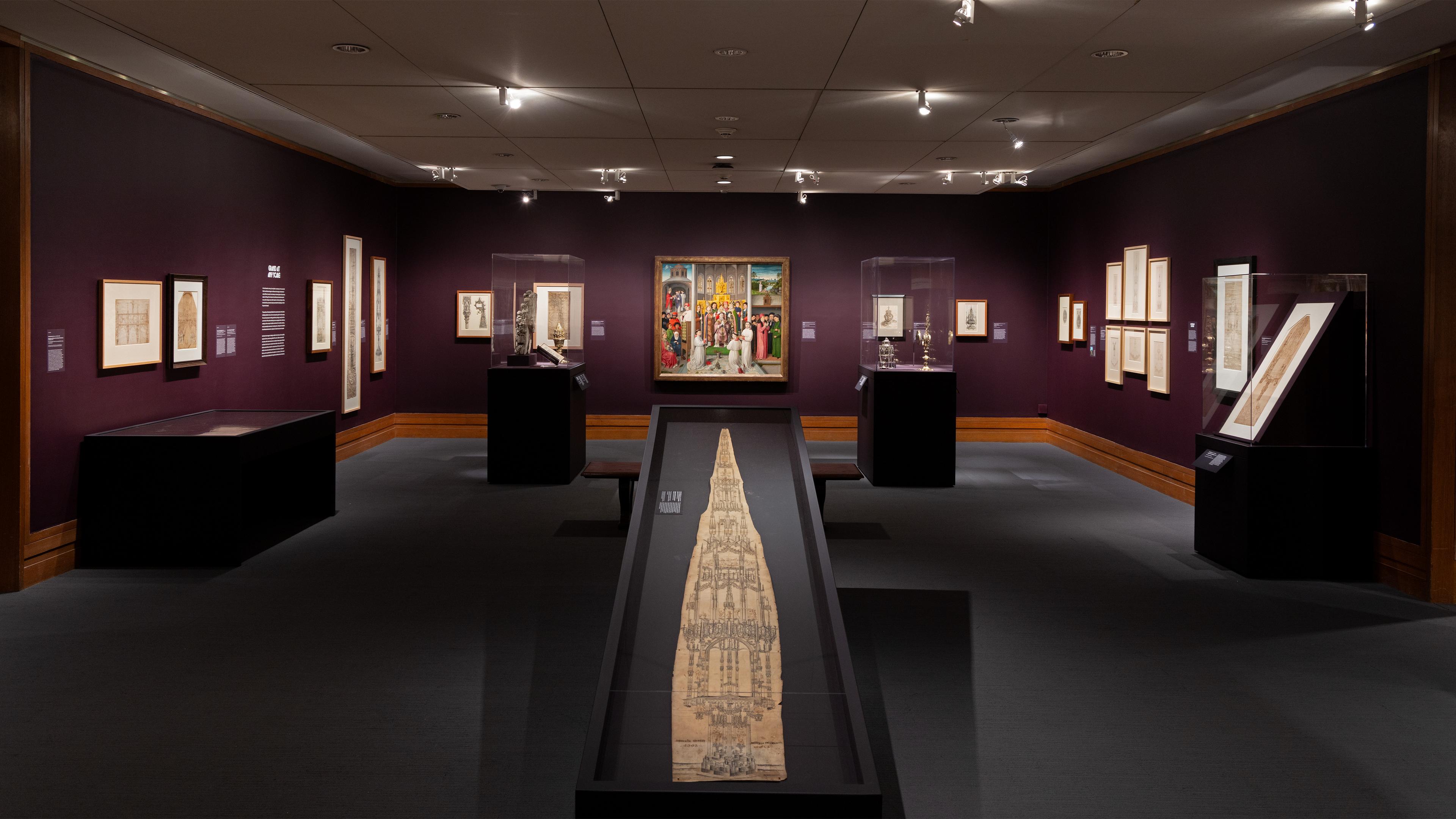 A dimly lit museum gallery with dark walls displays architectural drawings in frames and glass cases