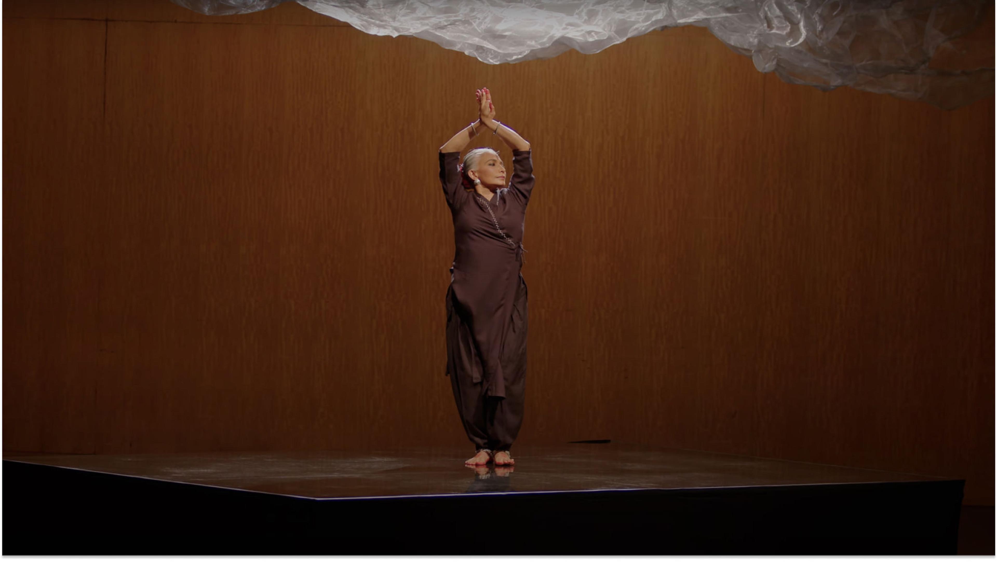 A woman stands on stage wearing a matching brown tunic and pants. Her arms are raised over her head with her palms pressed together.