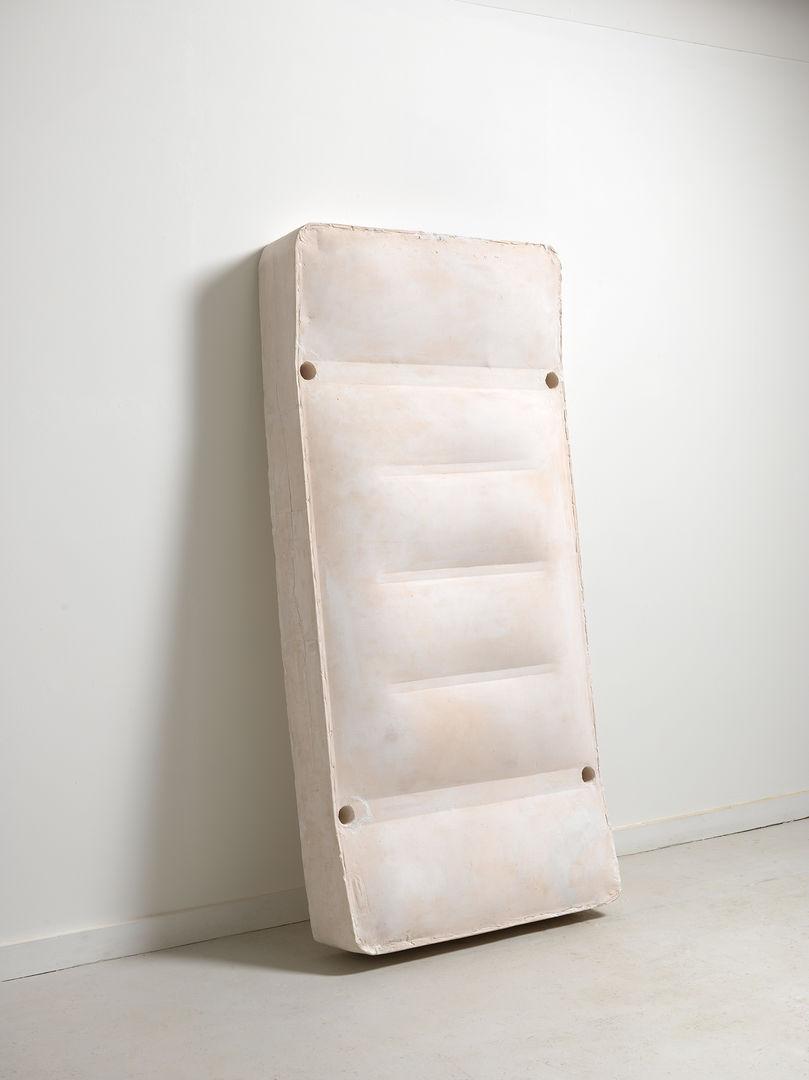 Sculpture of a white mattress set longways against a wall in a white space. The mattress bends gently and bears horizontal lines across the middle, suggesting slats from a bed frame.