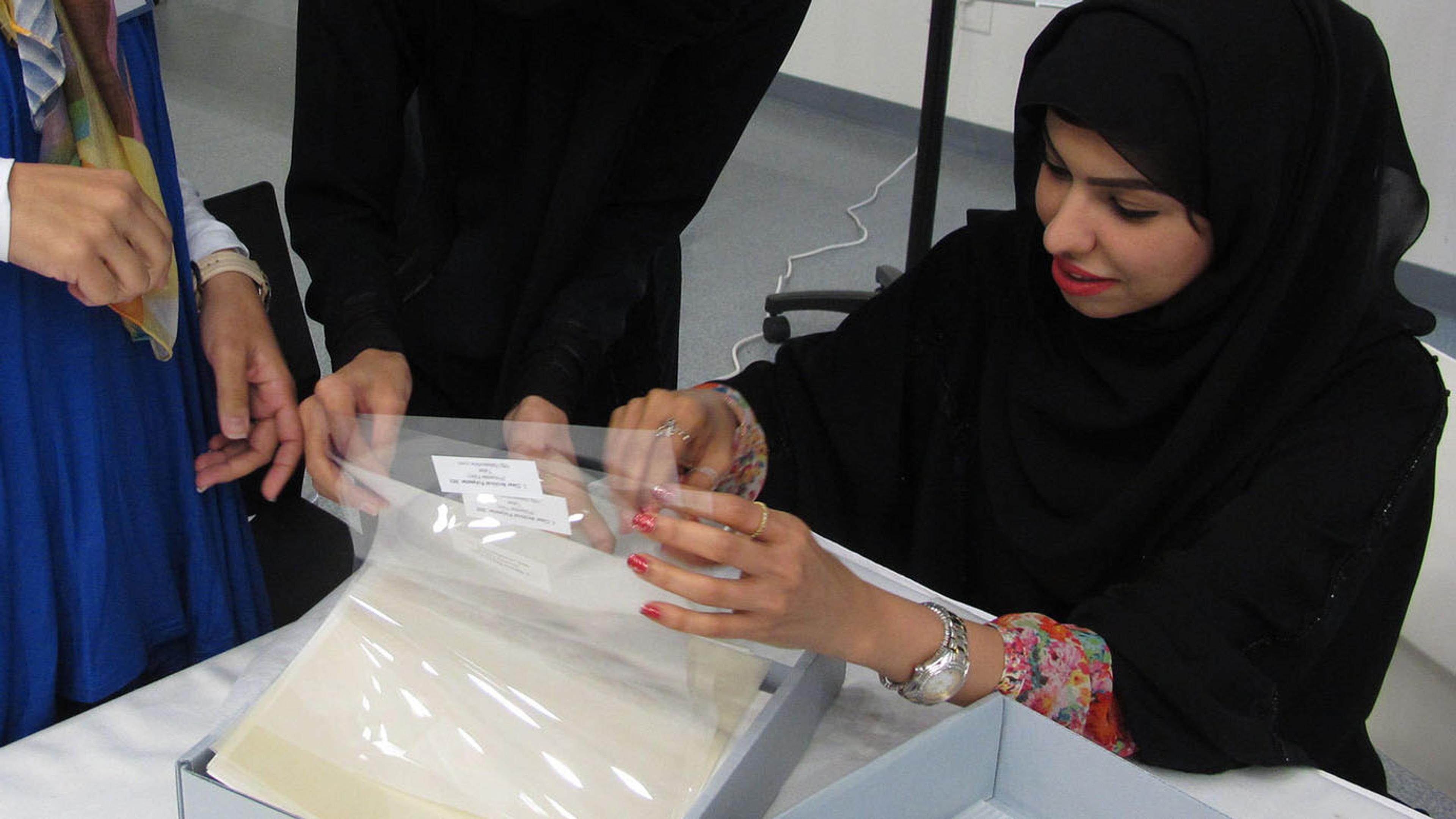 A group of women gather round a table to leaf through and consult papers protected in plastic sleeves stored in archival boxes.