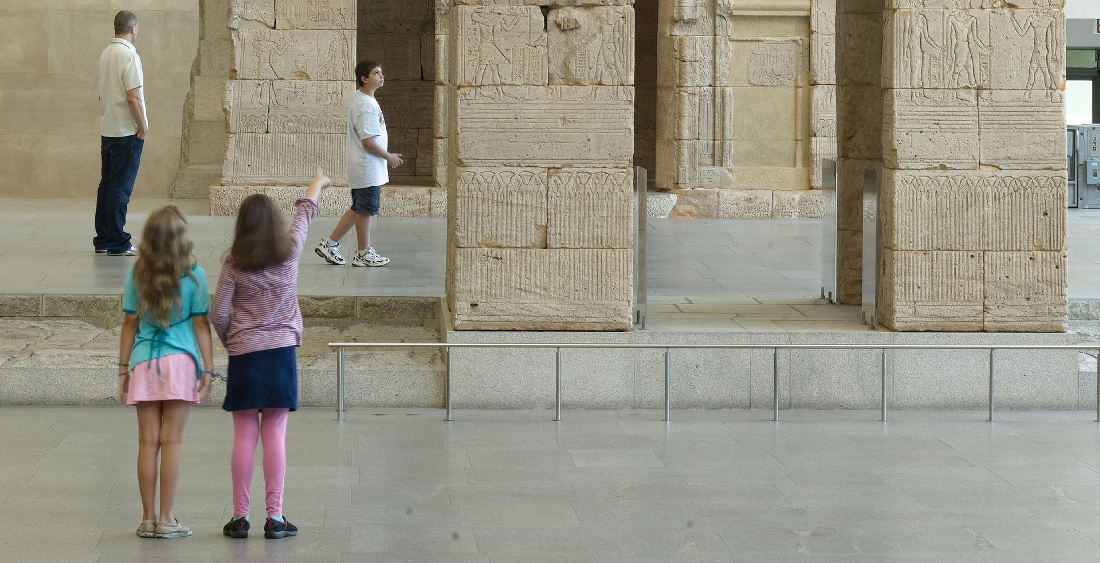 Two young children stand in front of the entrance of a stone Egyptian temple. The child on the right is pointing up to the structure.
