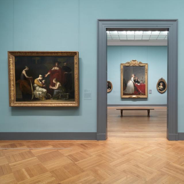 Art gallery interior featuring oil paintings of interior scenes in ornate gold frames, with a blue wall and wooden flooring.