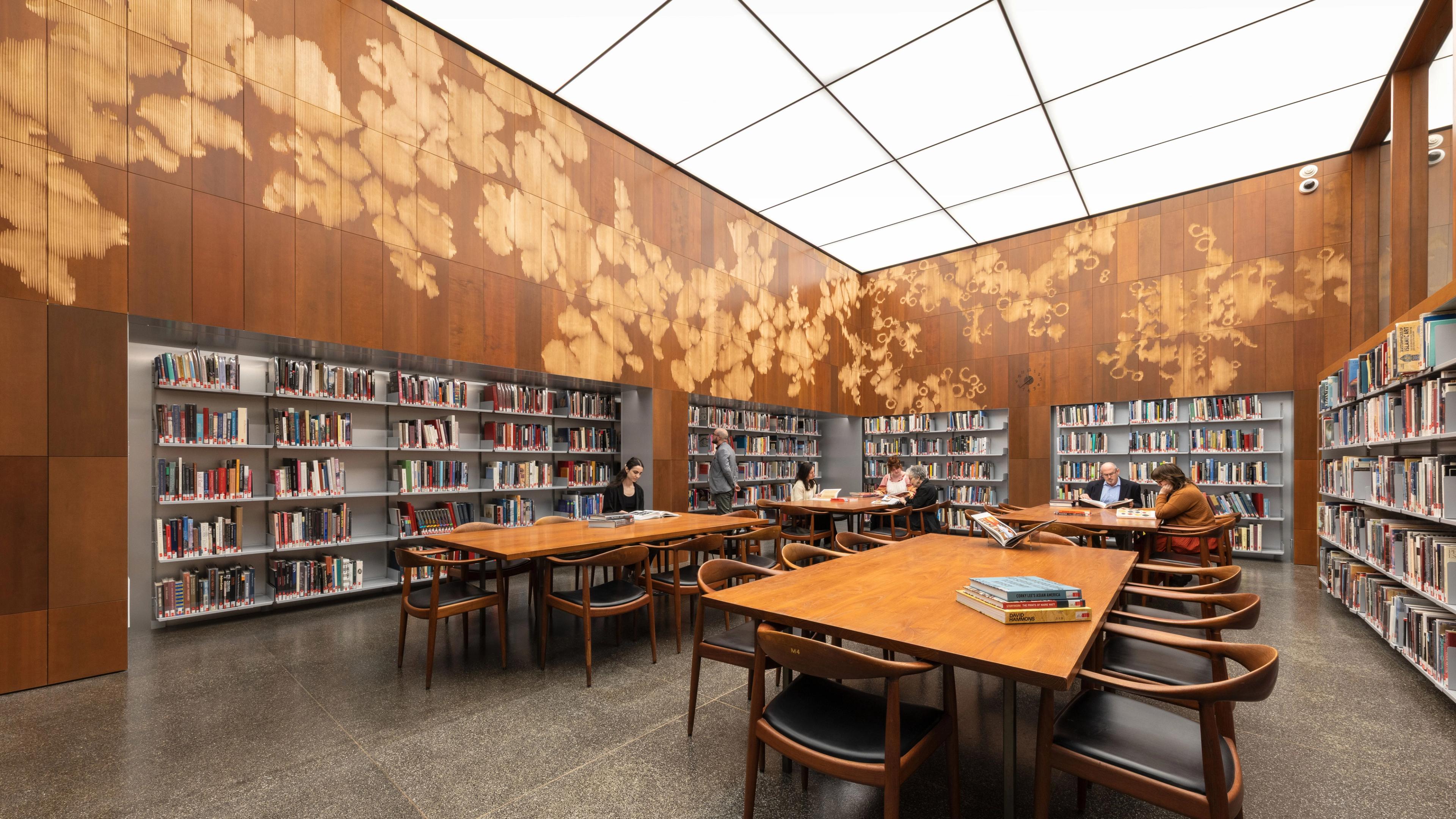 Brightly lit reading room with long tables, walls of book shelving, and wood-paneled walls with abstract motifs.