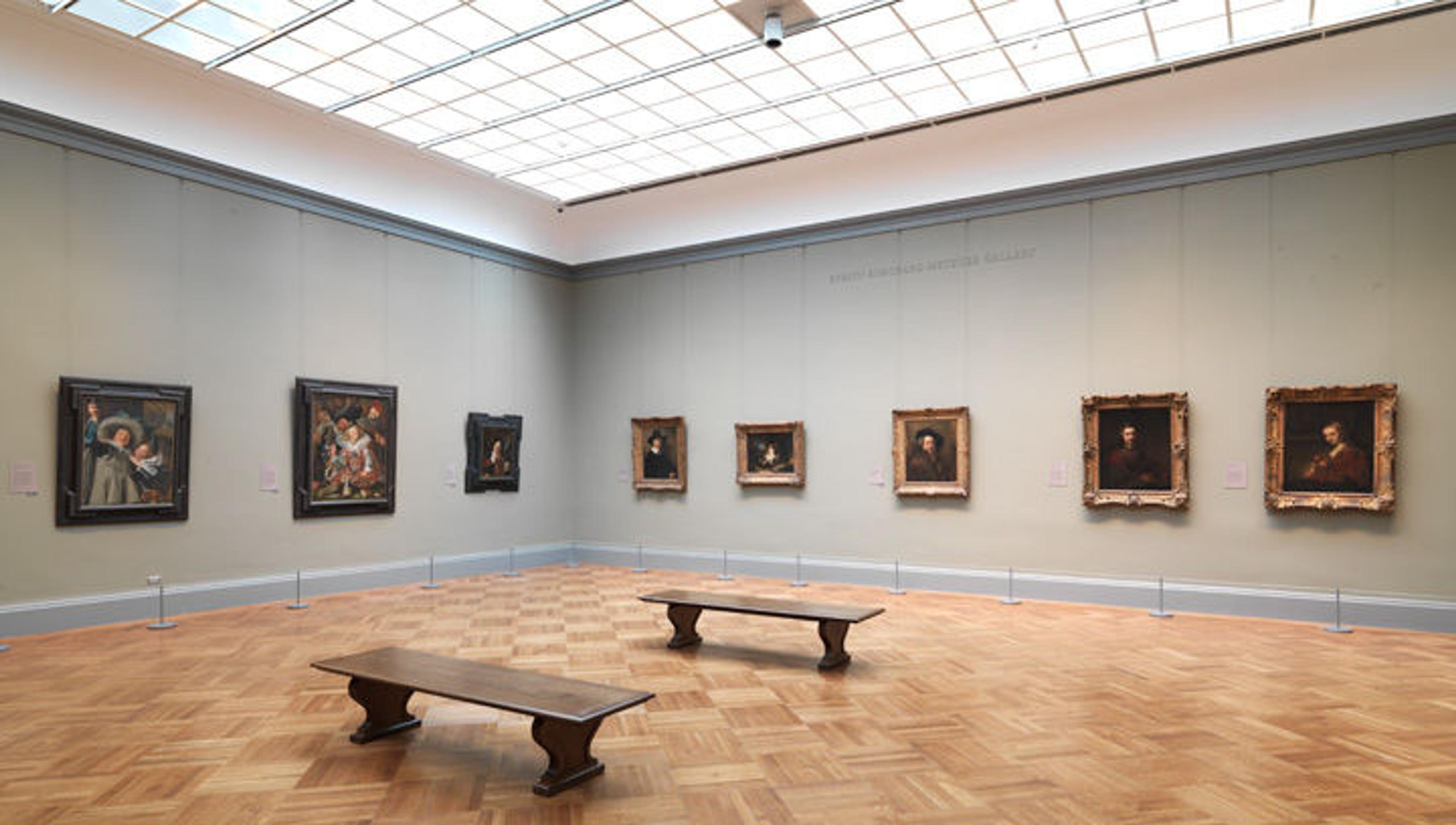 A spacious art gallery featuring oil paintings on the walls, two wooden benches, and a large skylight overhead. There are no people in the space.