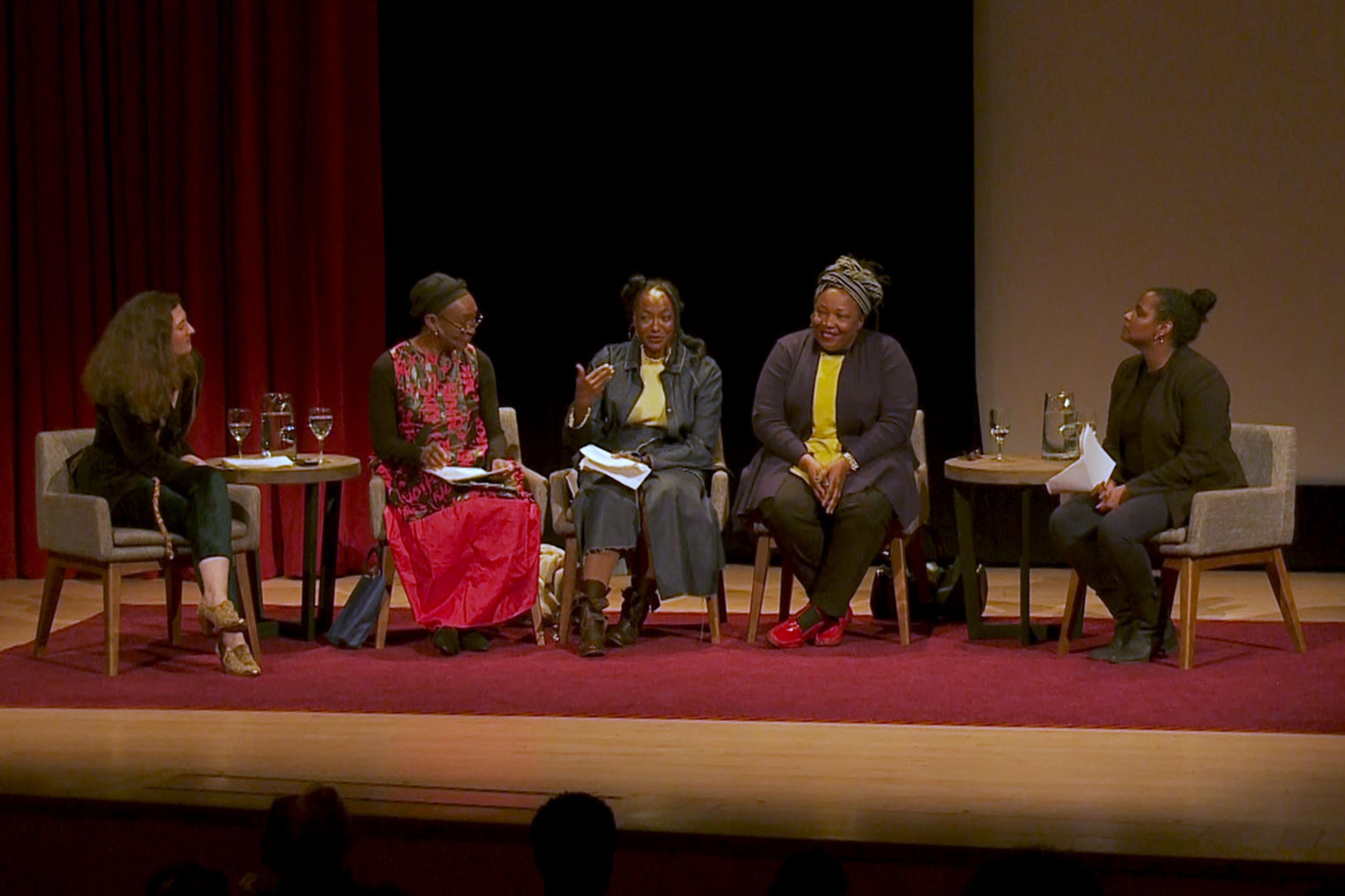 Five women are seated in a row under spotlights on a stage