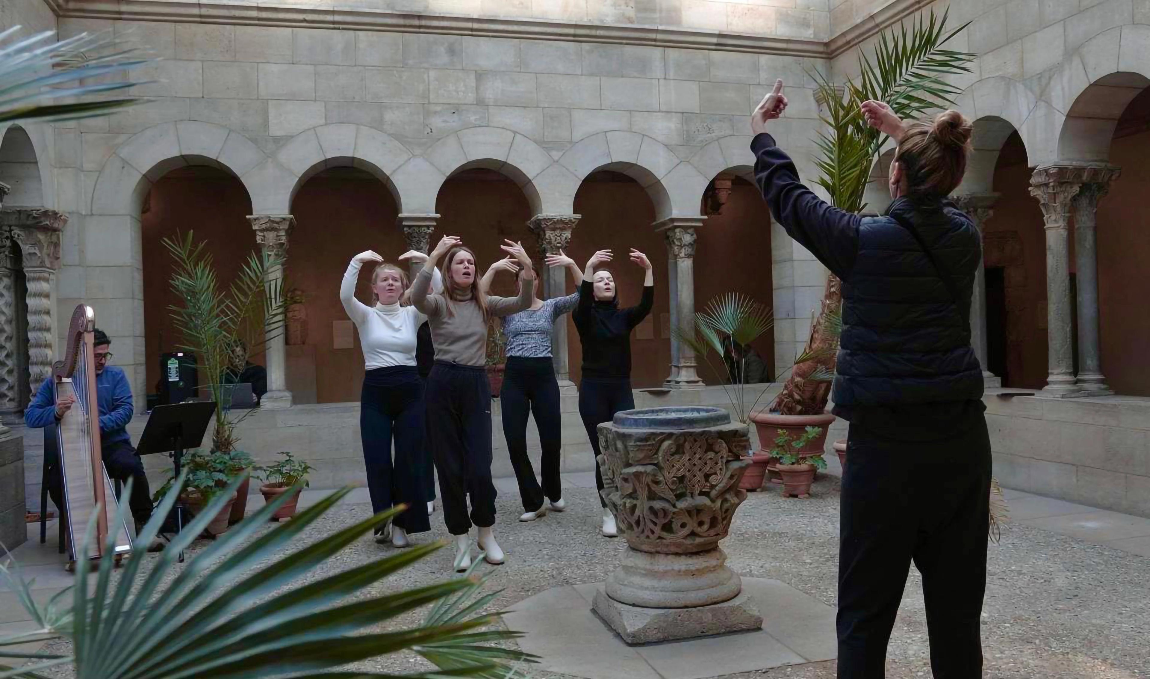 Performers in rehearsal at The Met Cloisters. A conductor guides four singers and dancers, their arms all outstretched, while a musician at left plays the harp.